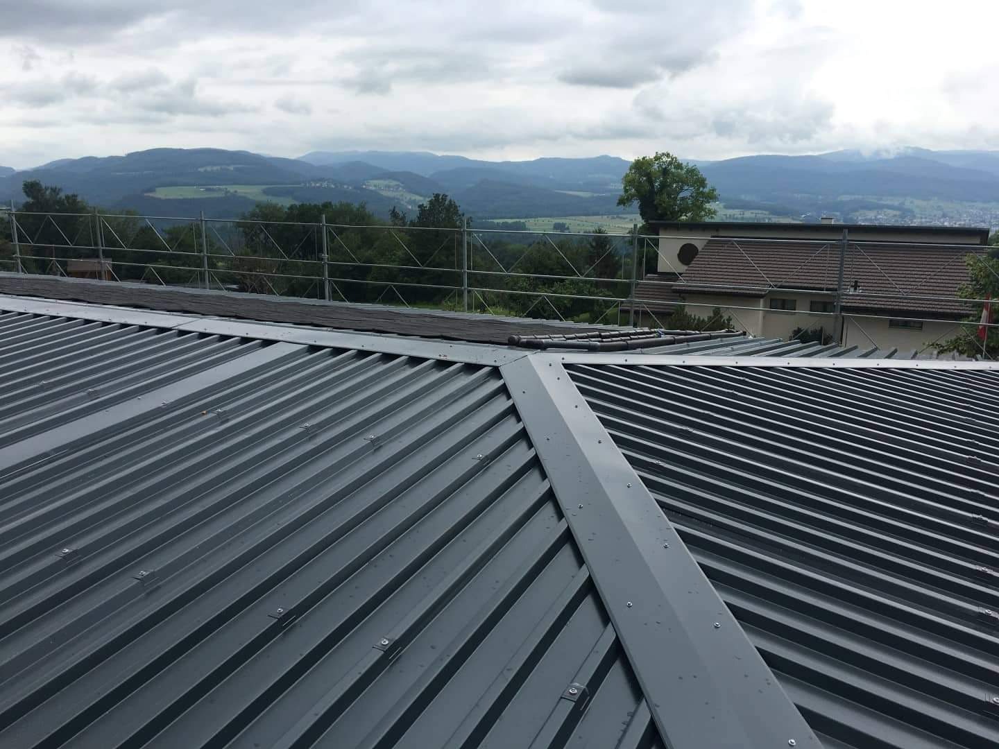 Szary dach z blachy trapezowej z widocznym systemem mocowań na tle krajobrazu górskiego i częściowo widocznych rusztowań.