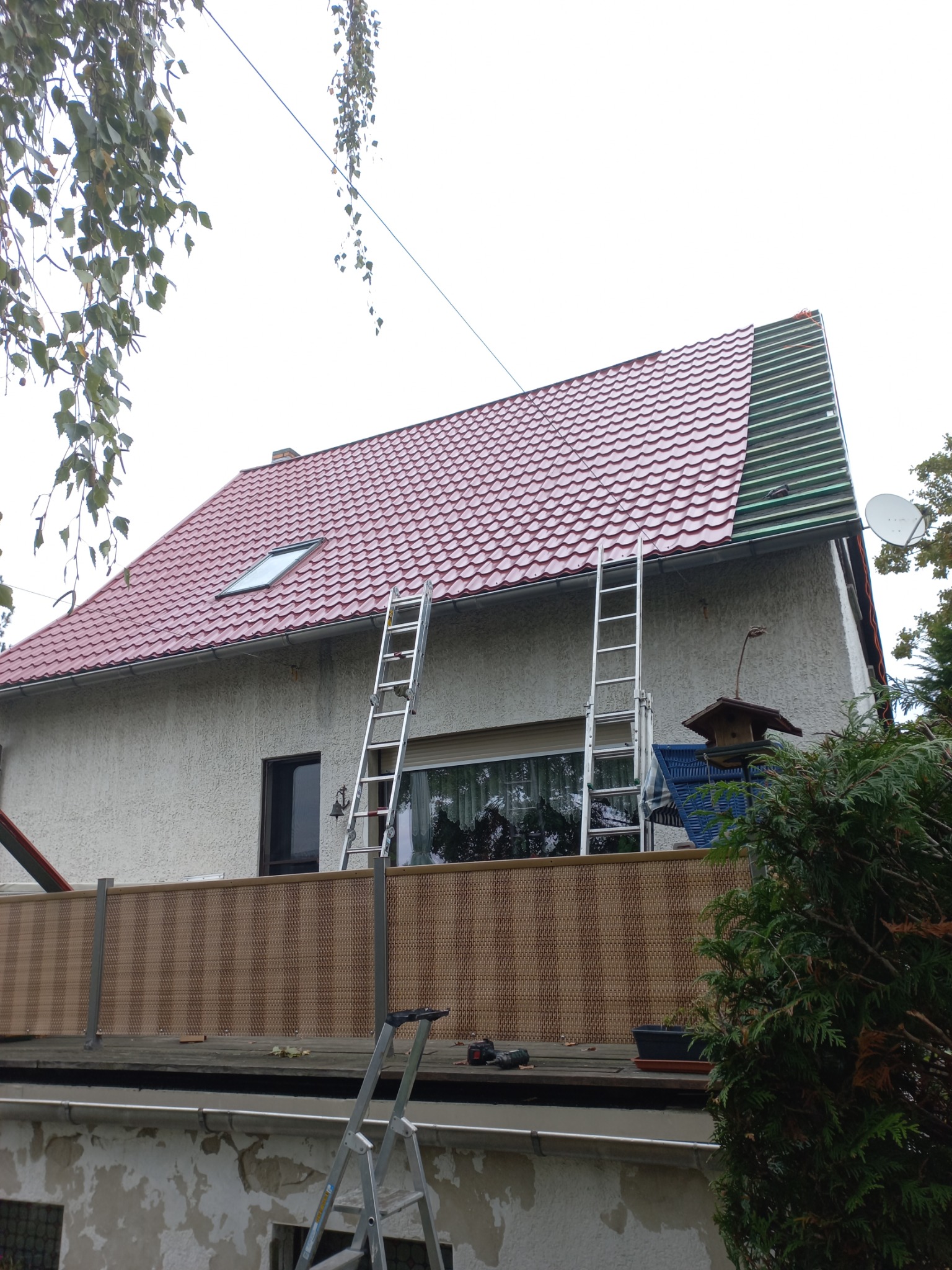 Dom z częściowo wymienionym czerwonym dachem, widoczne drabiny i fragment zielonego pokrycia dachowego w trakcie prac.