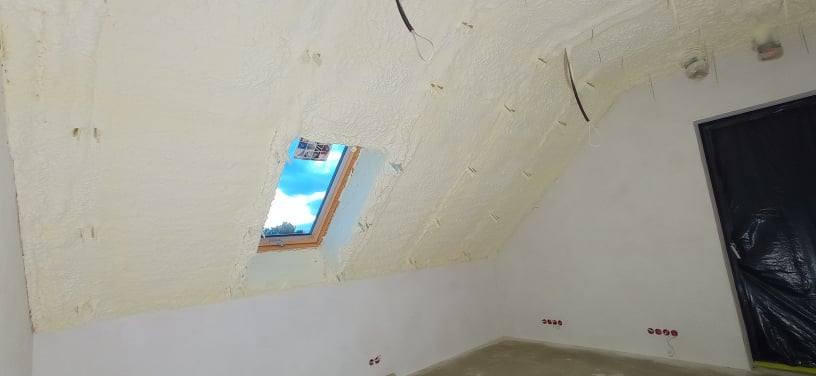 Poddasze po izolacji pianką PUR, widoczne okno dachowe z błękitnym niebem, wystające przewody elektryczne i zabezpieczone folią drzwi.