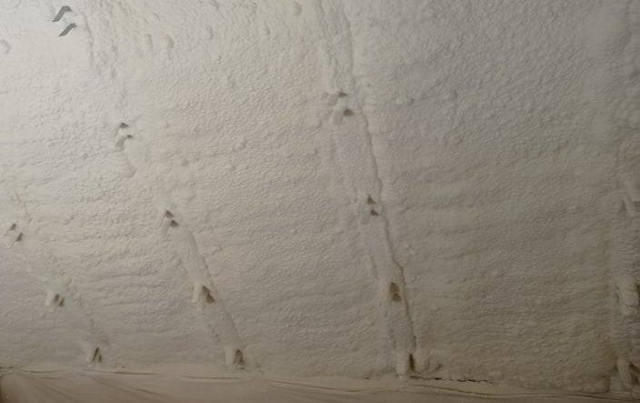 Poddasze pokryte grubą warstwą białej pianki izolacyjnej, widoczne szpary między krokwiami wypełnione pianą, nierówna powierzchnia izolacji.