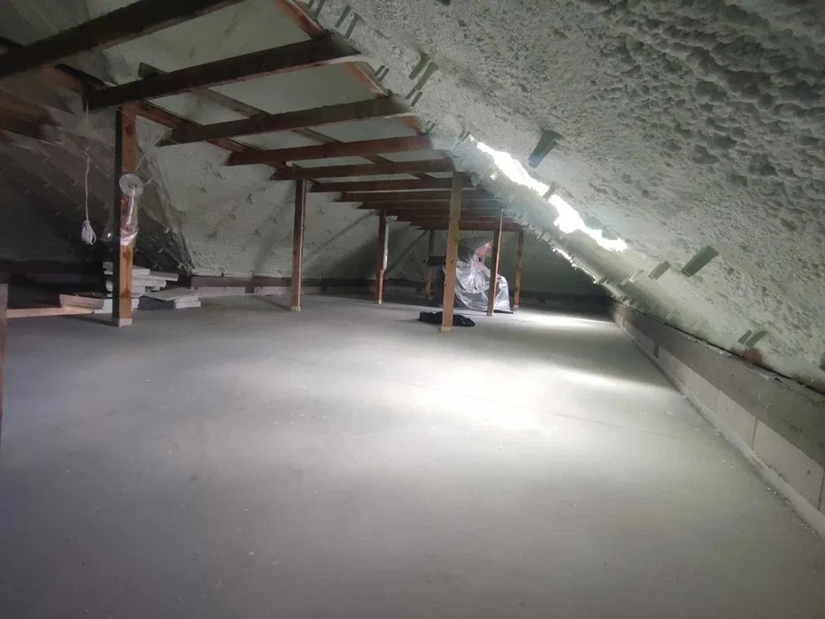 Poddasze po ociepleniu pianką PUR, widoczne drewniane elementy konstrukcyjne i biała pianka na skosach dachu, fragment podłogi z betonu.
