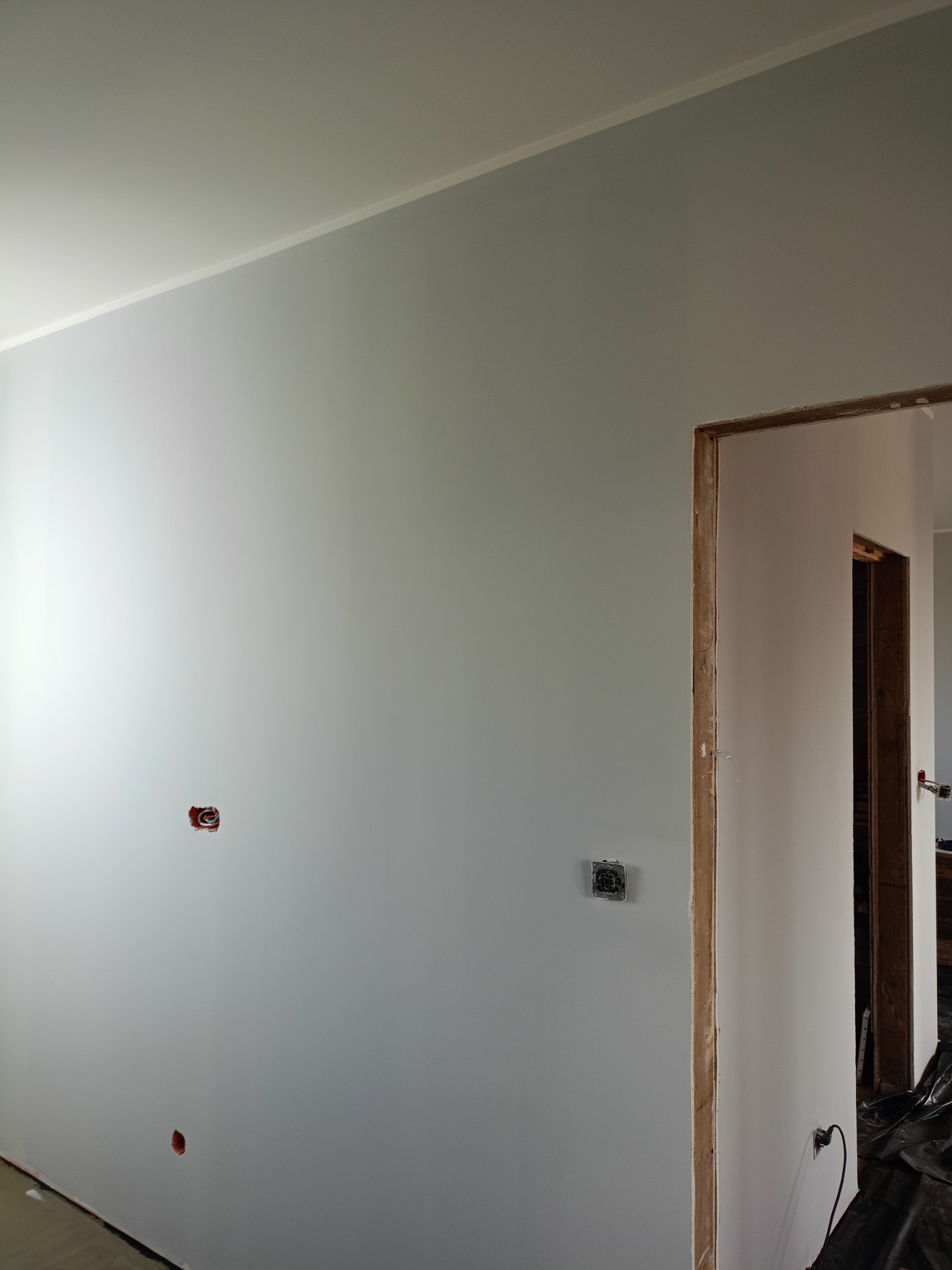 Ściana po malowaniu na szaro z widocznymi puszkami elektrycznymi i obramowaniem ościeżnicy drzwiowej. Widoczne niedoskonałości wykończenia.