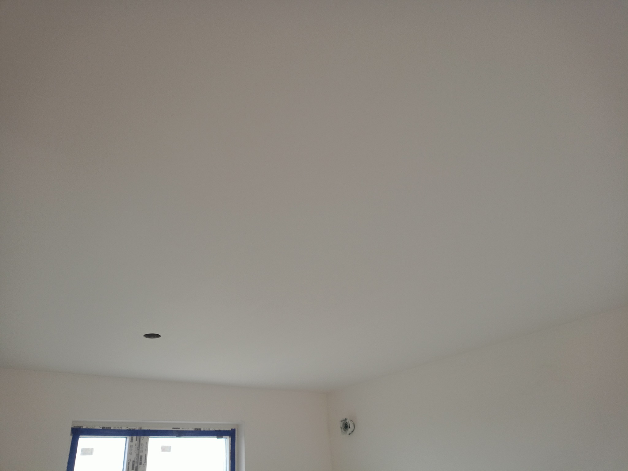 Białe, gładkie ściany i sufit w pokoju po malowaniu, z widocznym otworem na oświetlenie w suficie i oknem zabezpieczonym taśmą malarską.