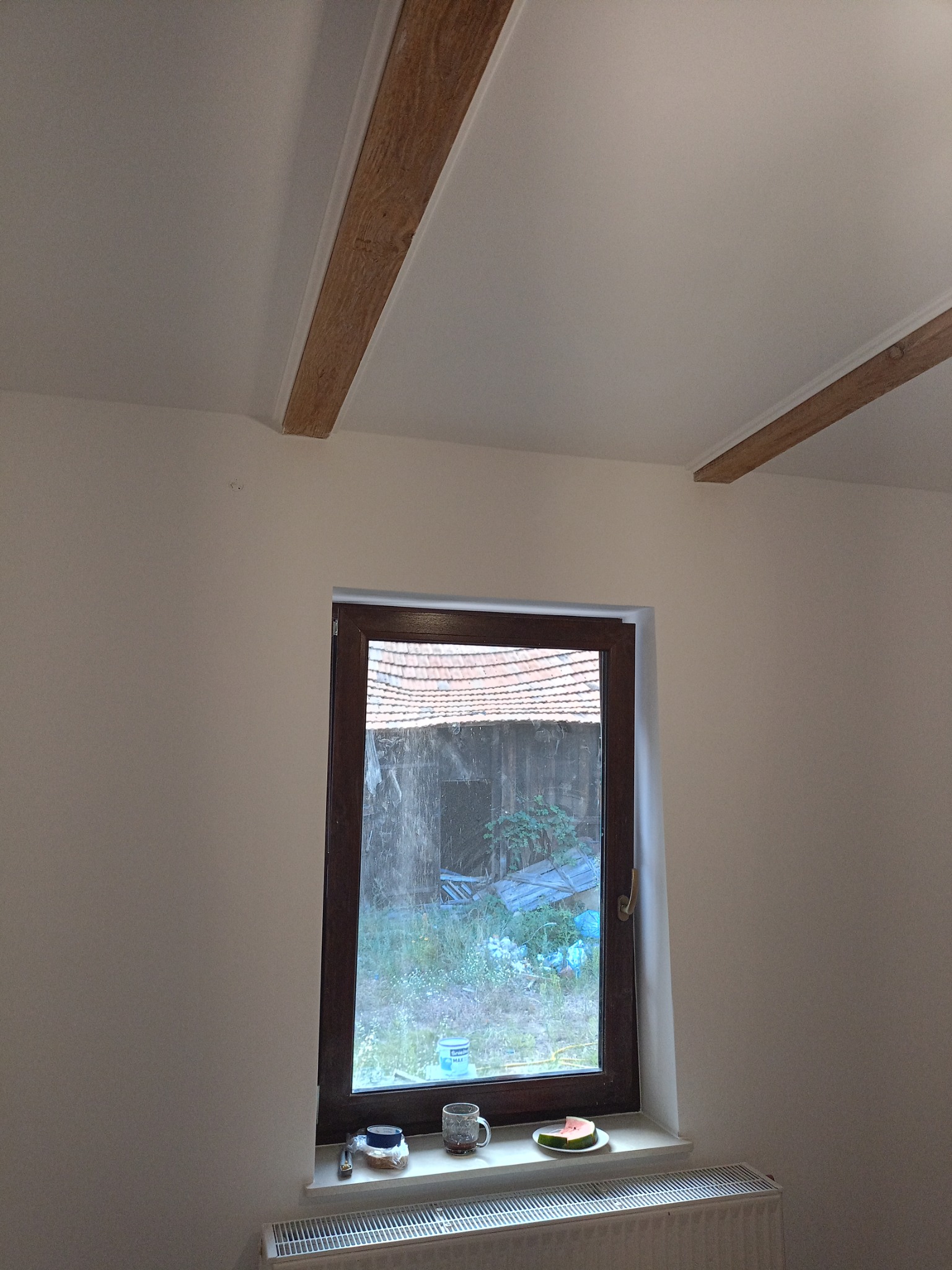 Jasne wnętrze po malowaniu z drewnianymi belkami stropowymi i oknem, na parapecie leżą przedmioty i arbuz. Widok na zewnątrz. Grzejnik pod oknem.