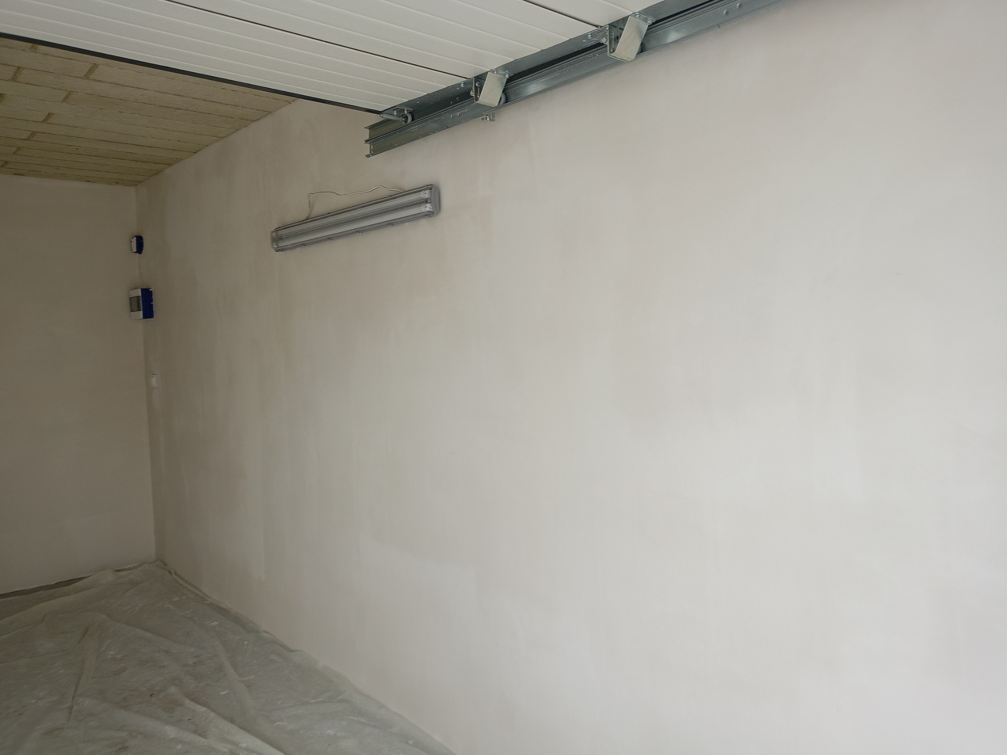 Wnętrze po szpachlowaniu: gładkie ściany w jasnym odcieniu, zabezpieczona folią podłoga, widoczne metalowe szyny sufitowe i lampa. Przygotowanie do malowania.