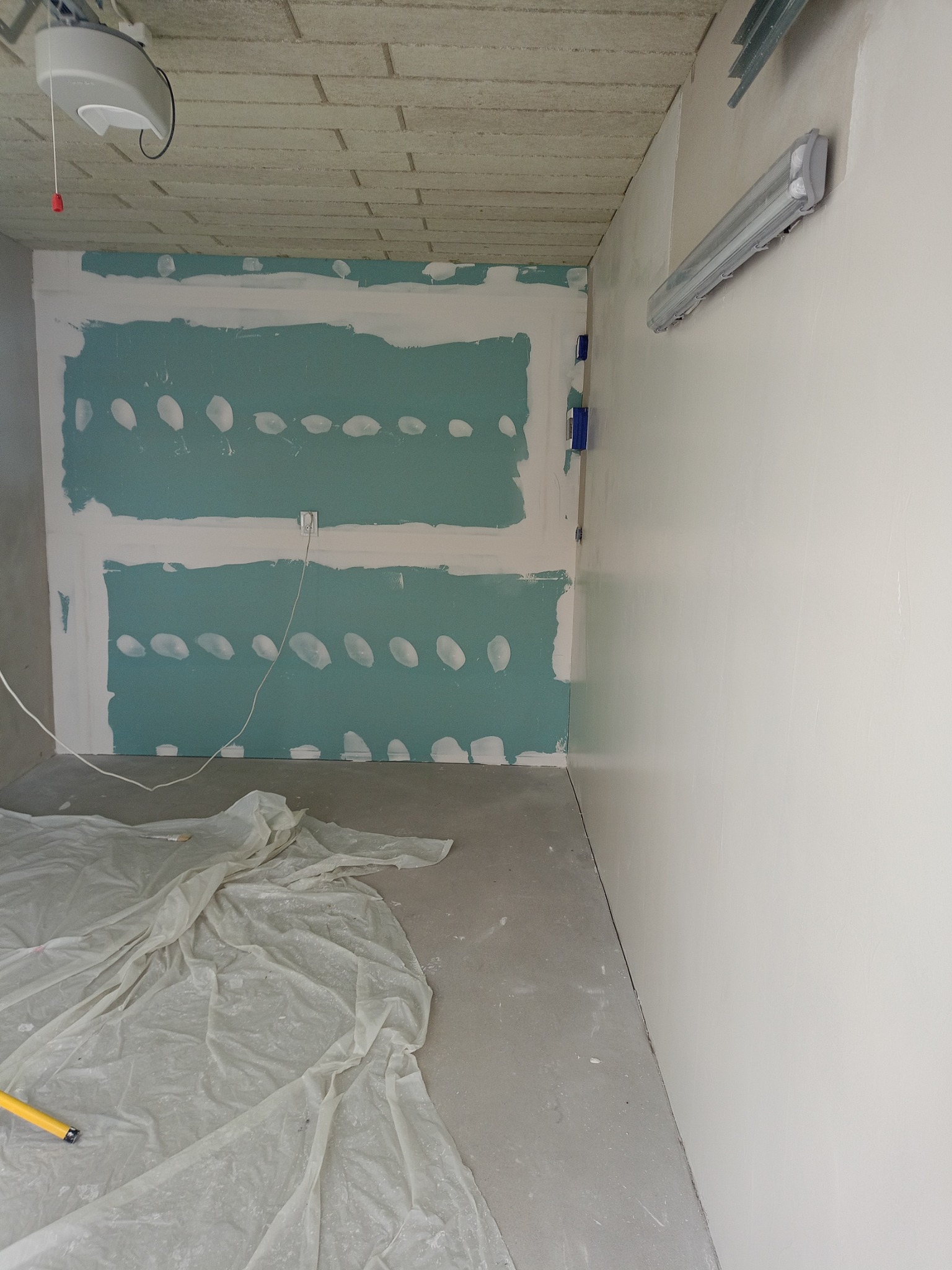 Ściana z płyt karton-gips, częściowo pokryta szpachlą, z widocznymi łączeniami. Podłoga zabezpieczona folią malarską. W tle surowy betonowy sufit.