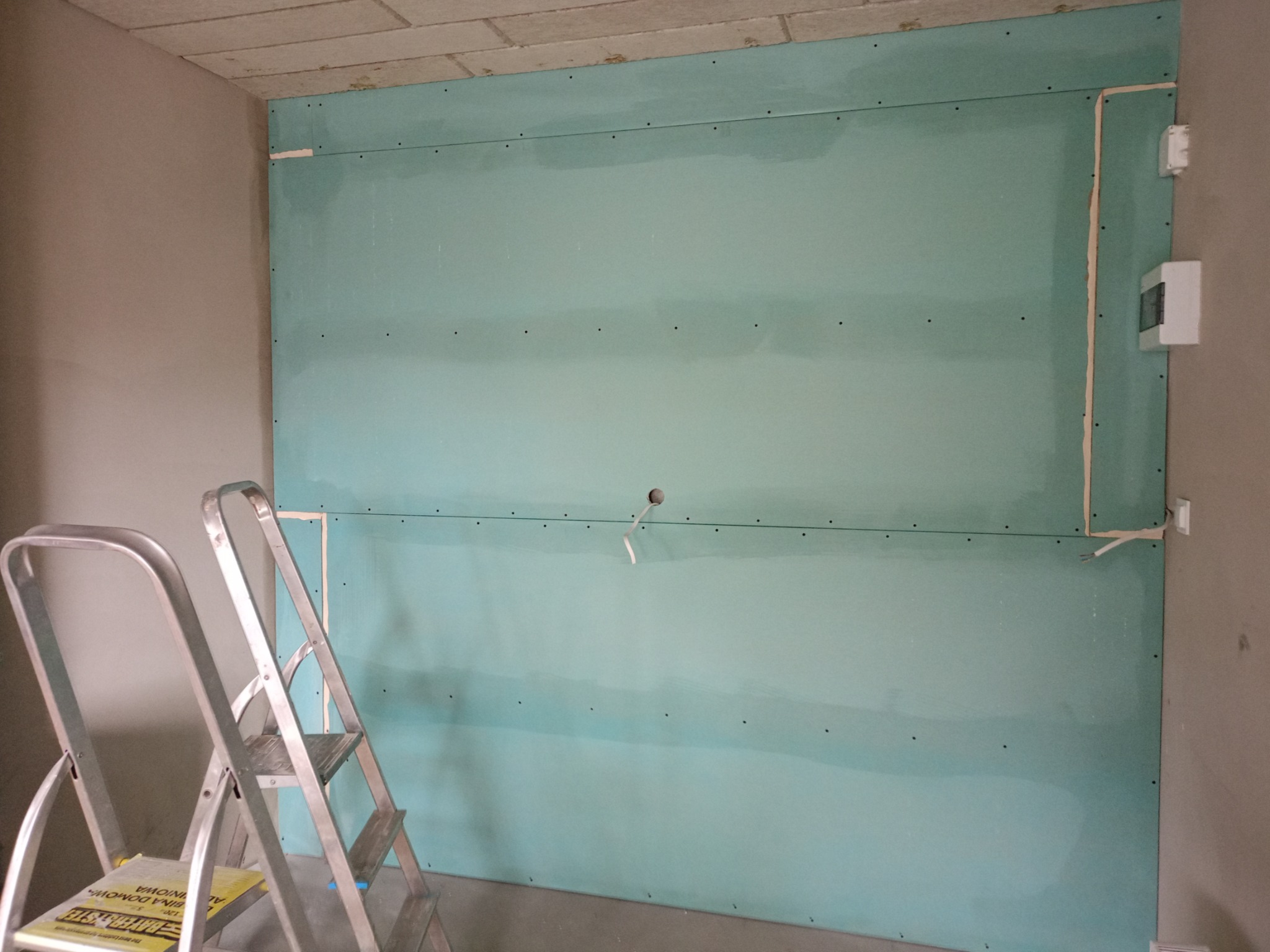 Ściana z zielonej płyty gipsowo-kartonowej, widoczne śruby mocujące, fragment instalacji elektrycznej i drabina aluminiowa z boku. Wnętrze w trakcie prac wykończeniowych.