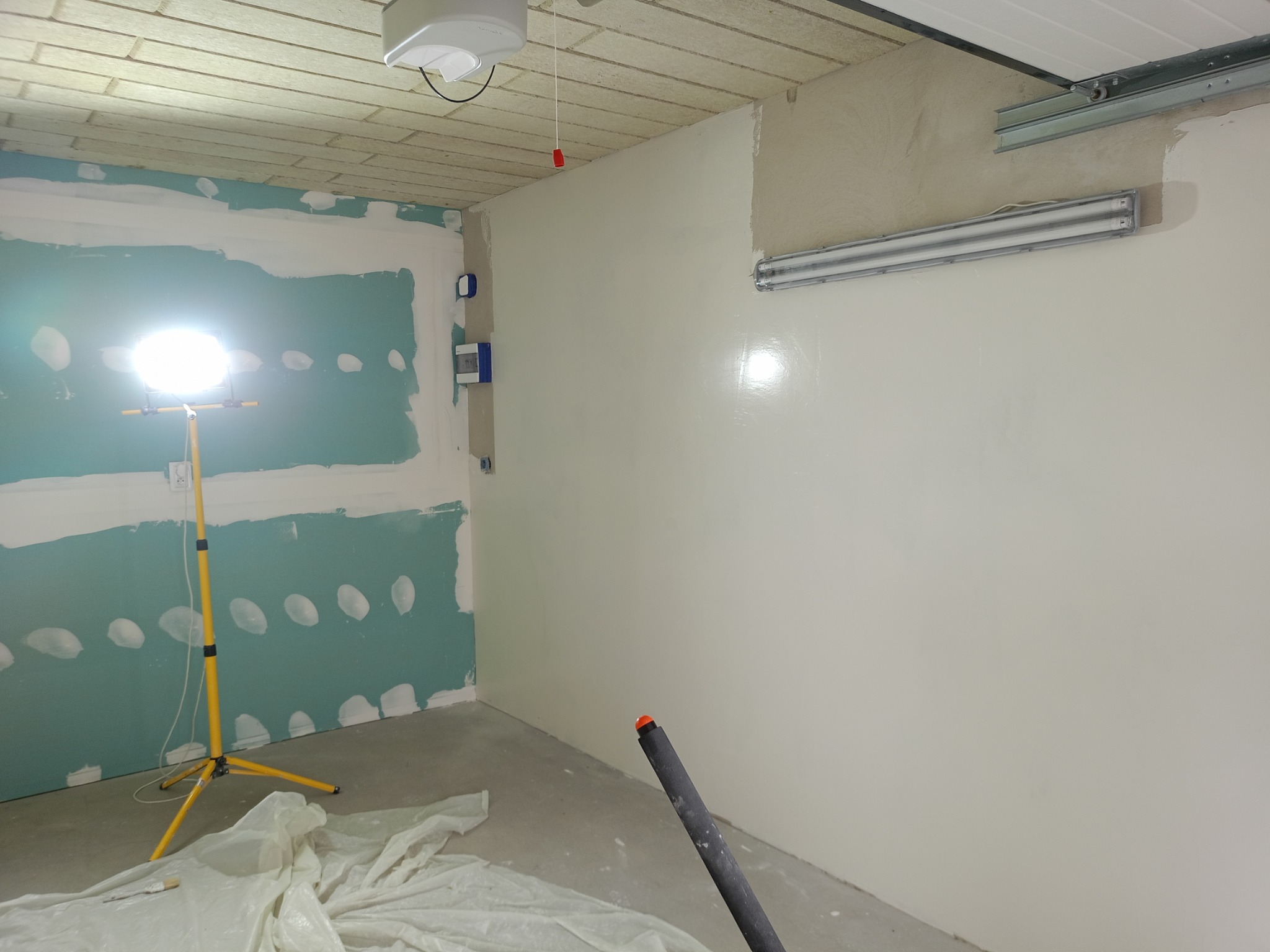 Ściana po szpachlowaniu i wstępnym malowaniu, widoczna lampa oświetleniowa i fragment ściany z płyt karton-gips. Częściowo zakryte podłogi folią malarską.