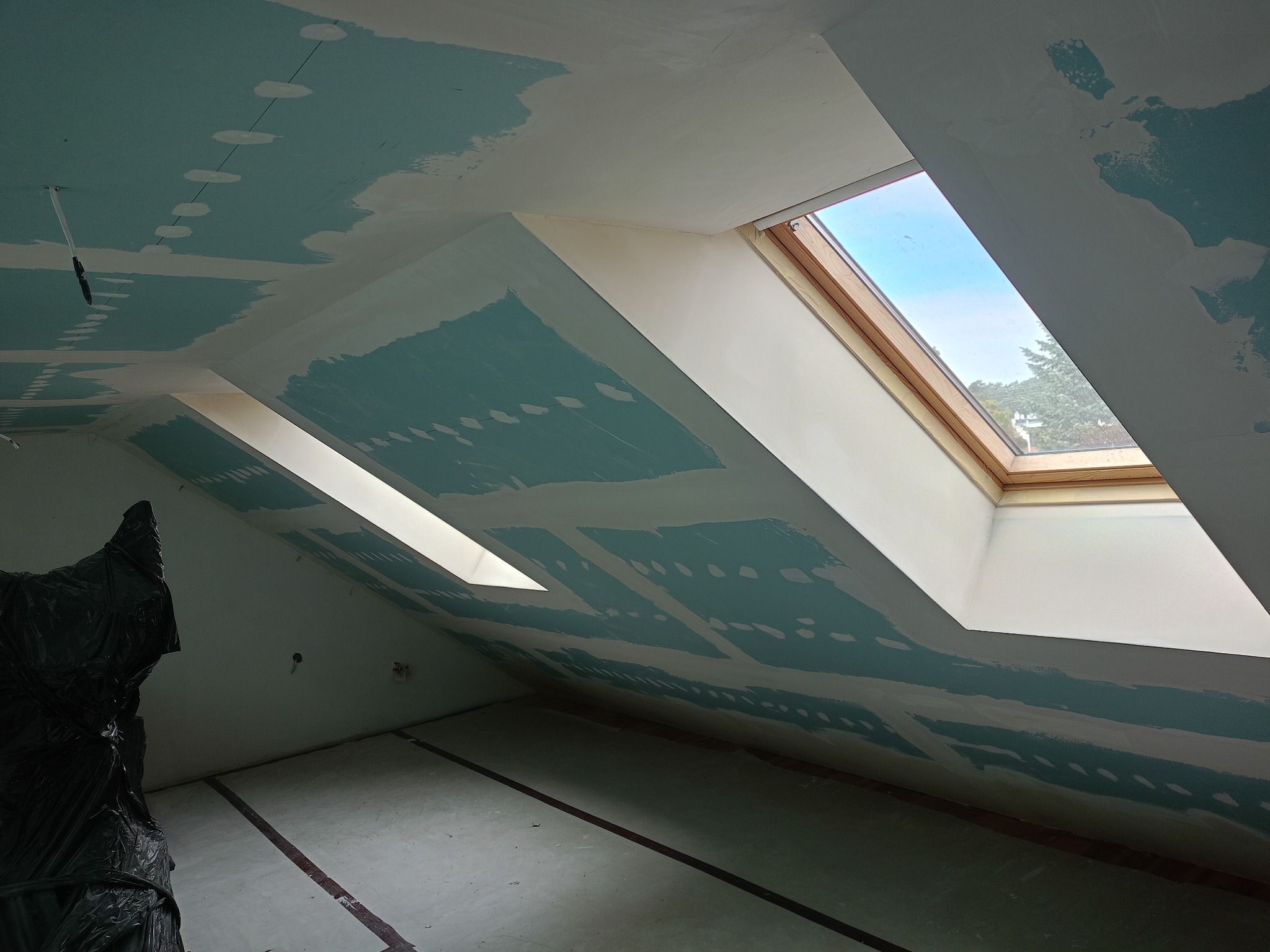 Poddasze ze skośnym sufitem w trakcie prac wykończeniowych; widoczne płyty karton-gips, szpachlowane łączenia i okna dachowe z widokiem na zewnątrz.
