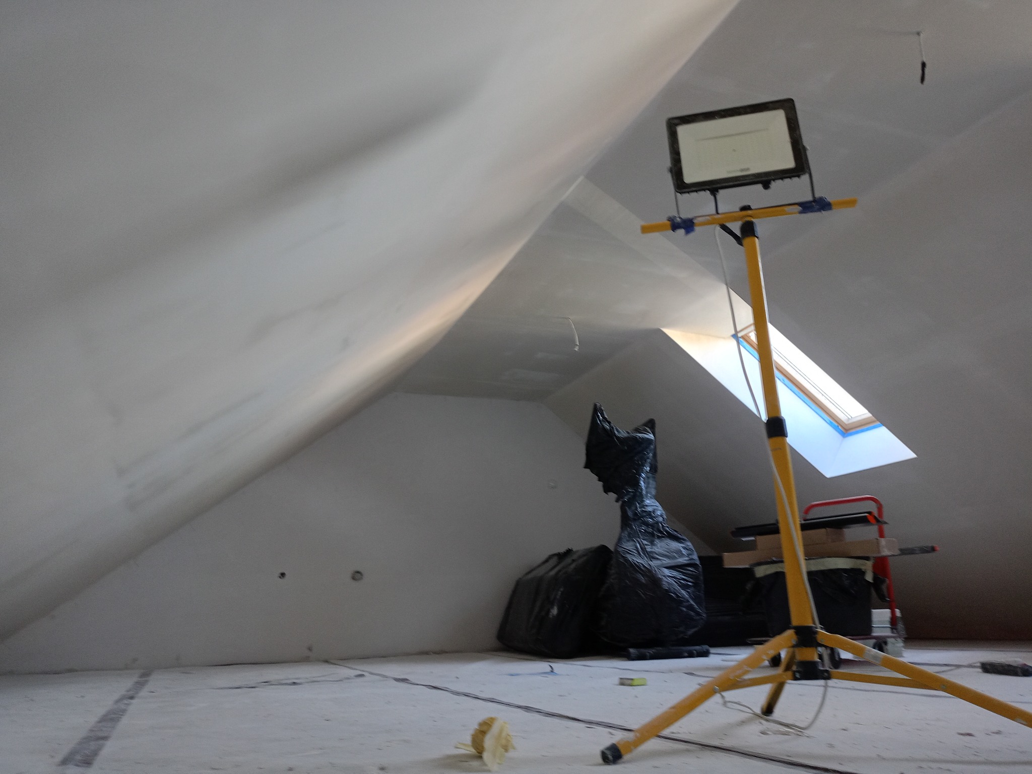 Poddasze ze skośnymi ścianami po szpachlowaniu, oświetlone halogenem na statywie. Widoczne worki z odpadami budowlanymi i okno dachowe.