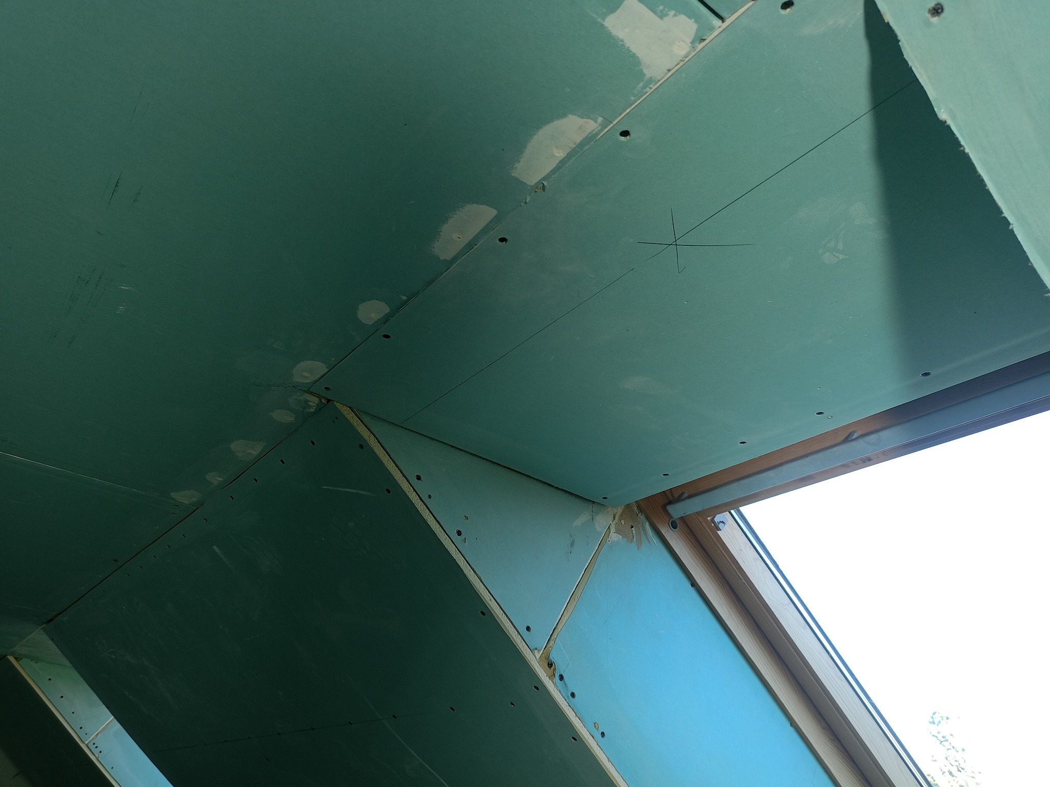 Montaż zielonej płyty gipsowo-kartonowej na poddaszu, widoczne ślady po szpachlowaniu i okno dachowe z drewnianą ramą. Ujęcie z dołu, perspektywa zbieżna.