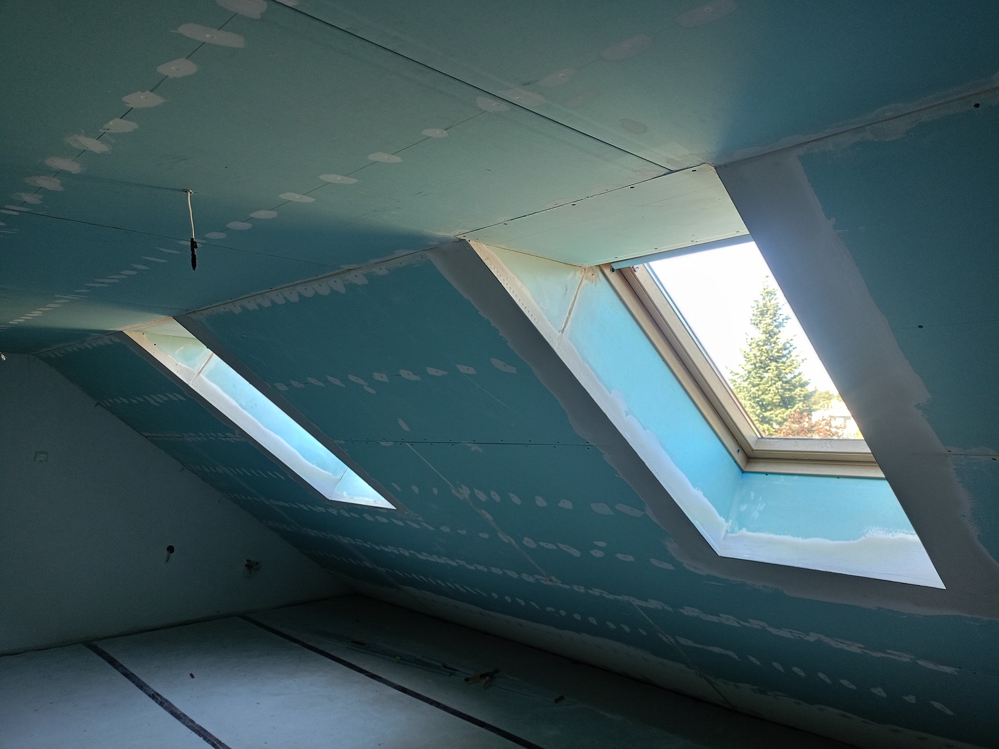 Poddasze z oknami dachowymi w trakcie wykańczania płytami gipsowo-kartonowymi. Widoczne łączenia płyt i szpachlowane fragmenty. W oknach widok na zewnątrz.