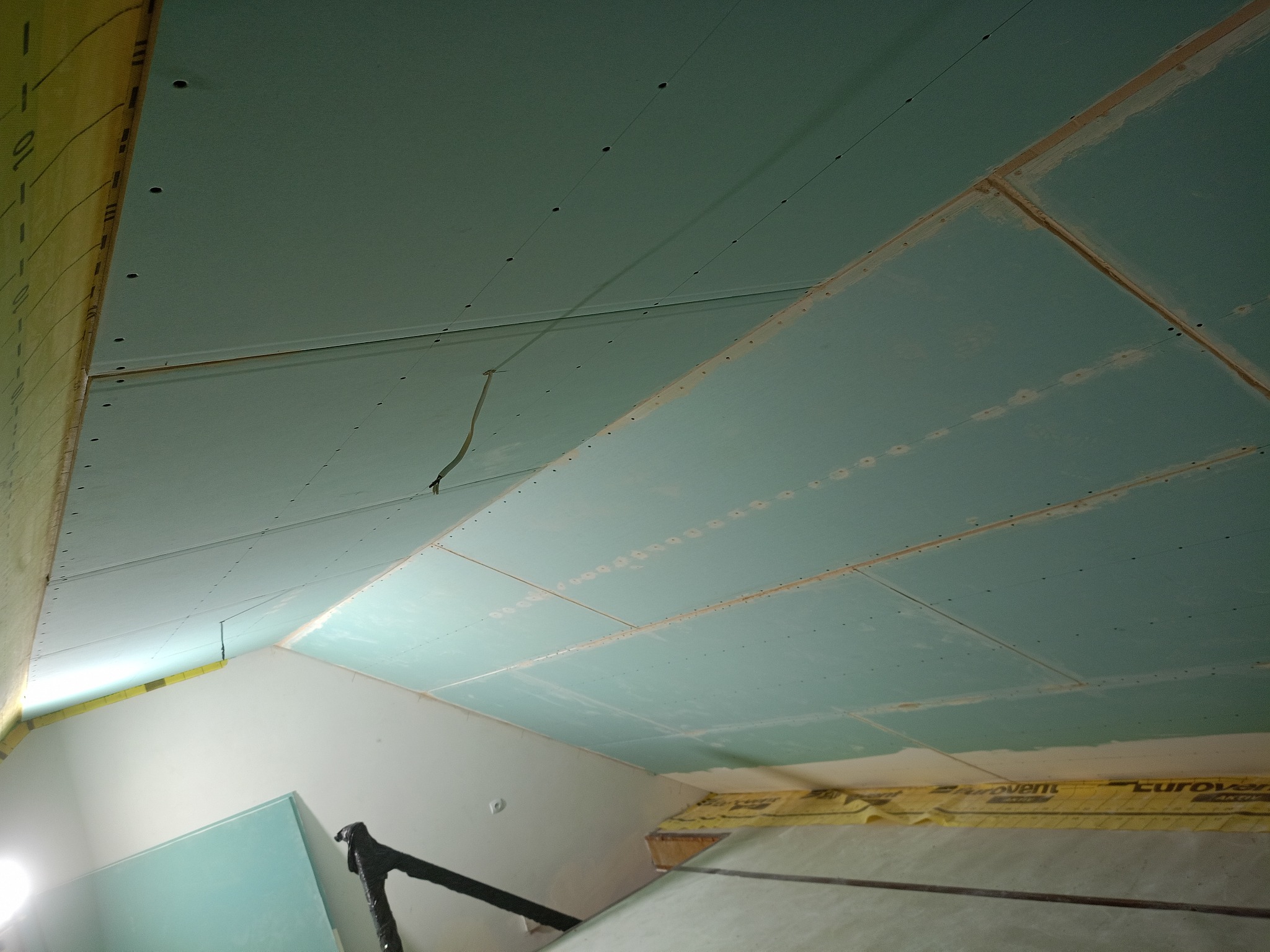 Skośny sufit poddasza z zamontowanymi płytami kartonowo-gipsowymi, widoczne łączenia płyt i przygotowane otwory na śruby. Przewody elektryczne wystają ze szczeliny.