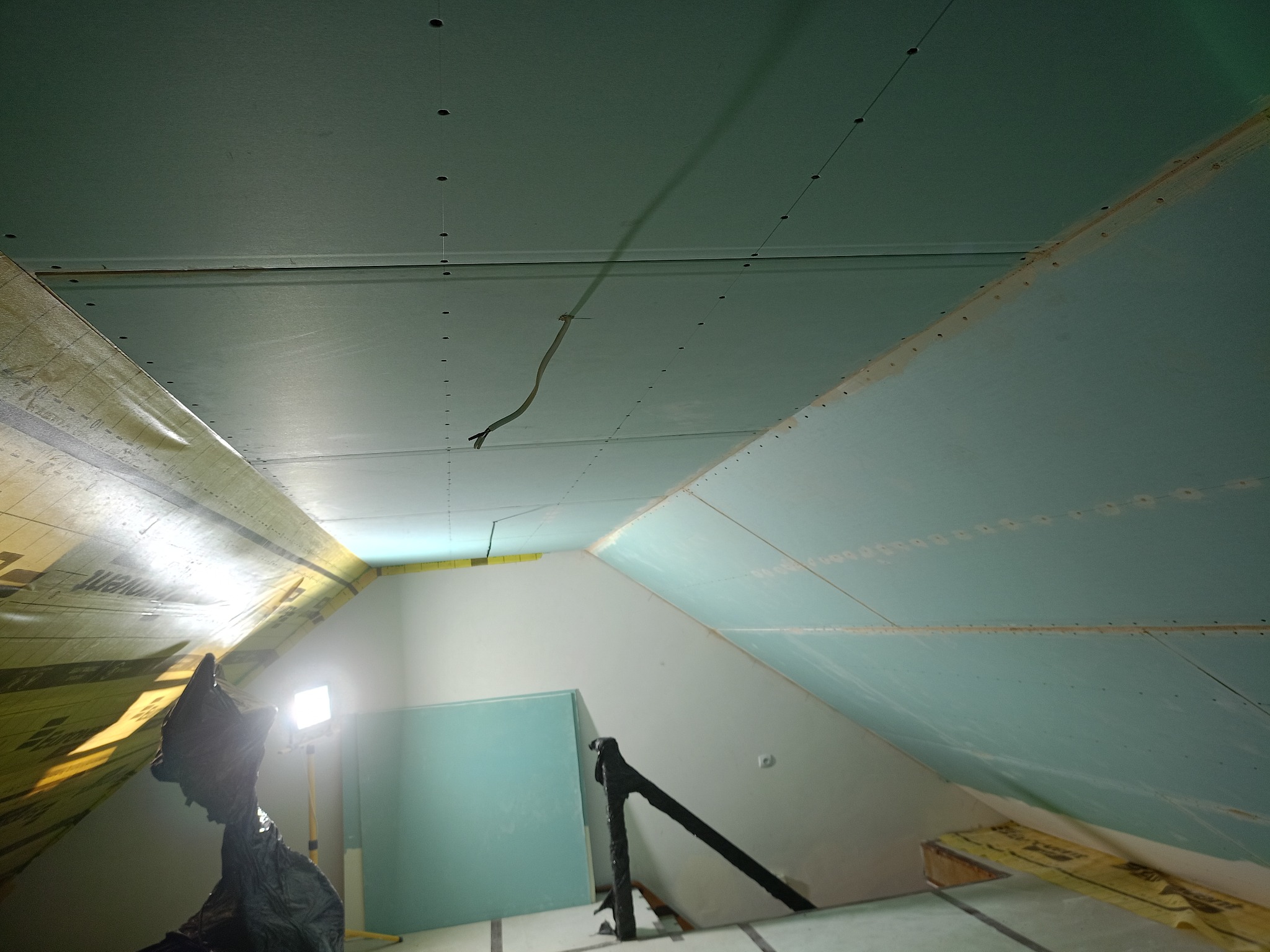 Montaż płyt karton-gips na poddaszu, widoczne mocowania i kable. Częściowo ocieplone, ukośny sufit. Praca w toku, widoczne materiały budowlane.