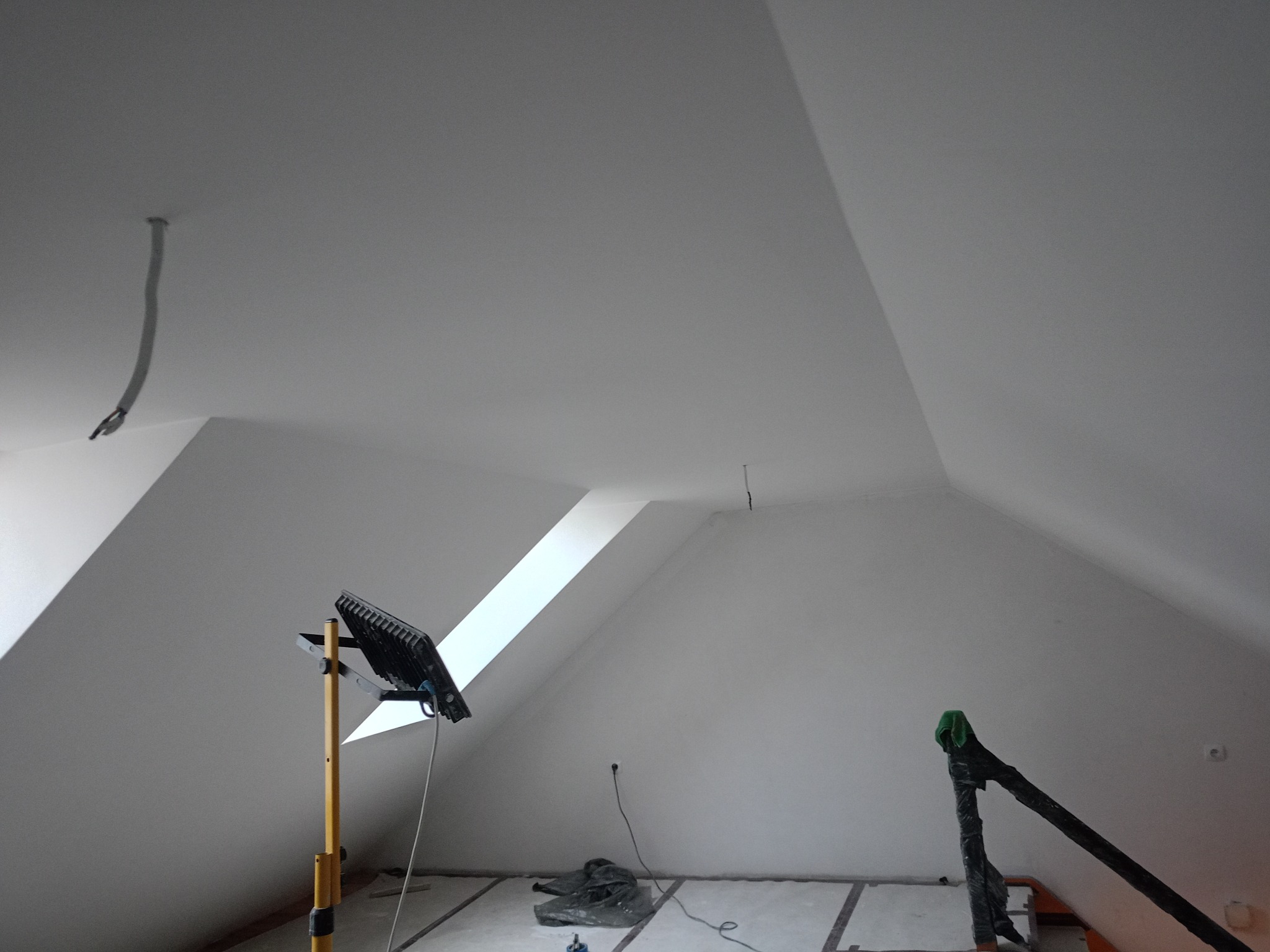 Białe poddasze po malowaniu z oknem dachowym, widoczne narzędzia malarskie i lampa oświetleniowa. Świeżo pomalowane ściany i sufit.