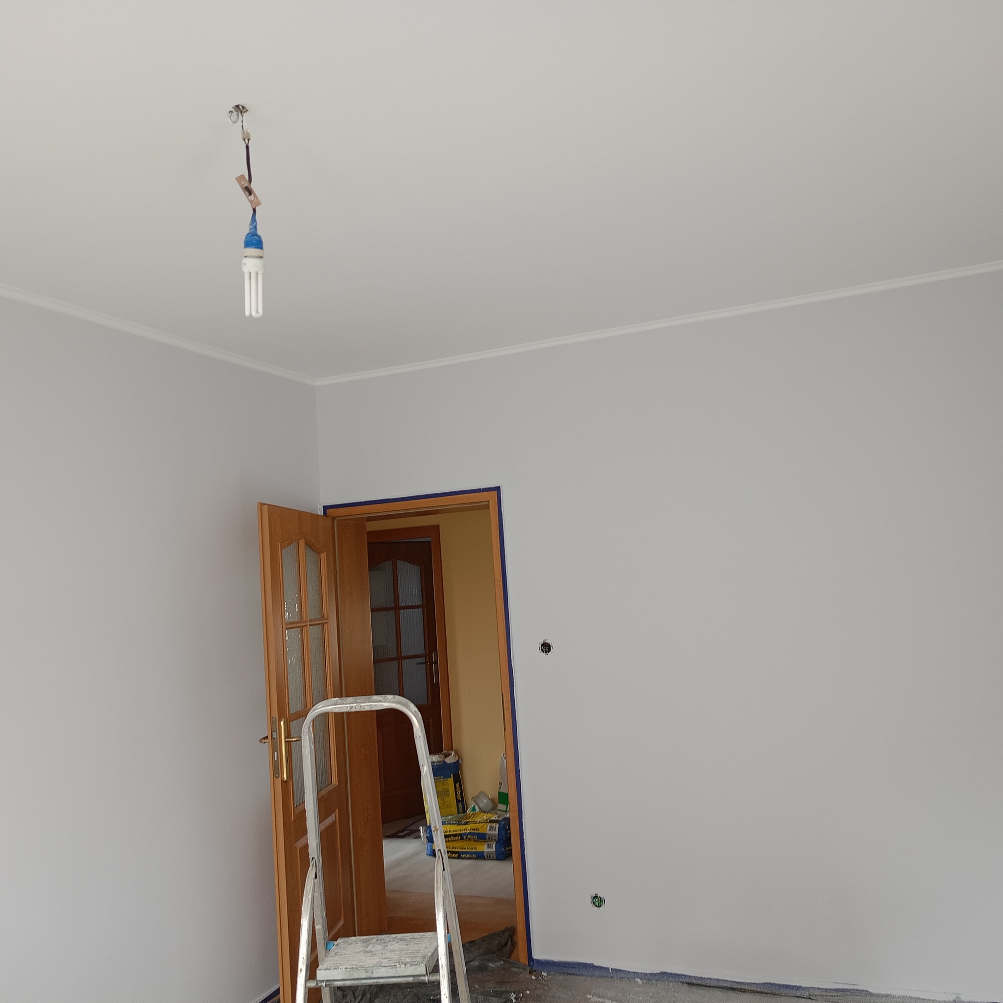 Wnętrze po malowaniu na szaro, widoczna drabina, otwarte drzwi z zabezpieczeniem taśmą malarską. Żarówka w suficie. Worki z zaprawą w tle.