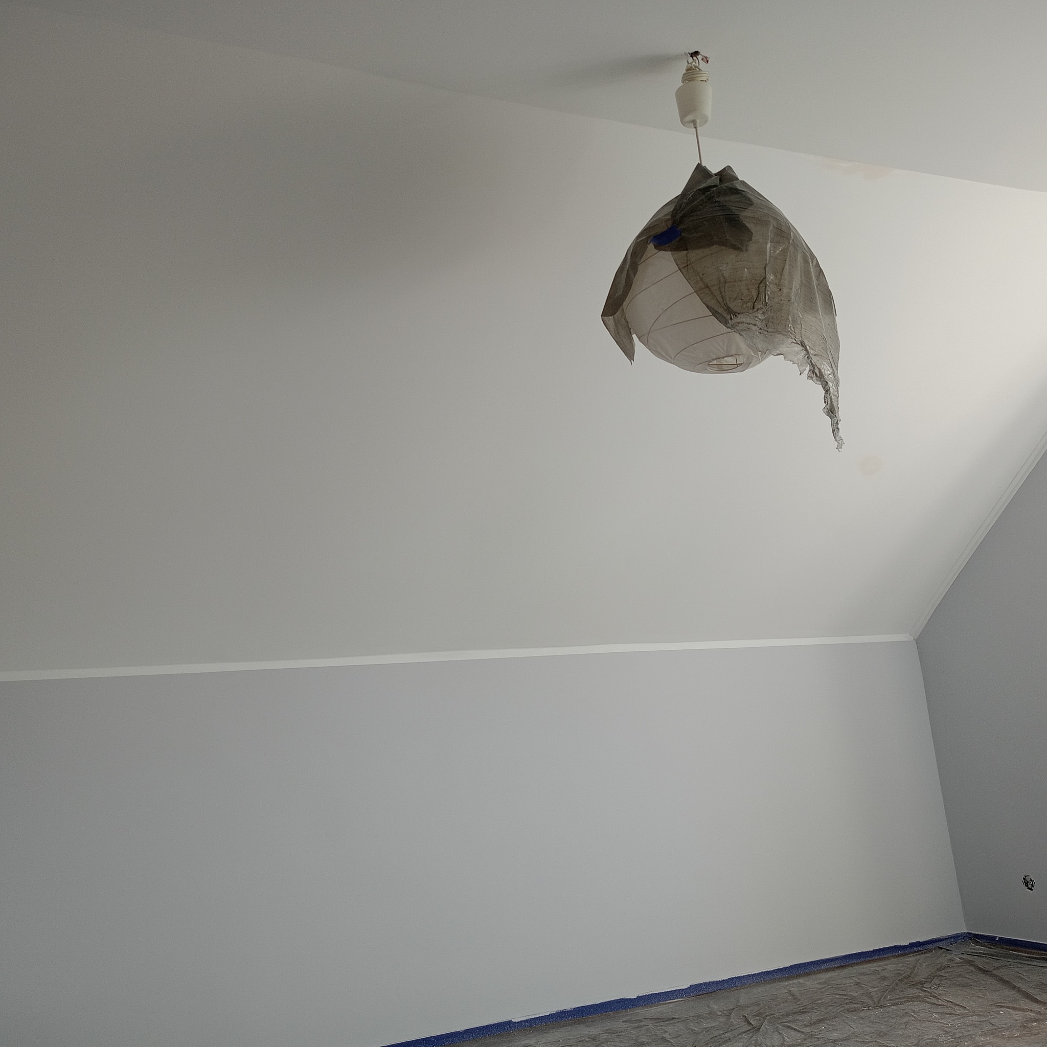 Świeżo pomalowane na szaro wnętrze pokoju z zabezpieczoną folią podłogą i lampą osłoniętą folią malarską. Widoczna listwa przypodłogowa i skos dachu.
