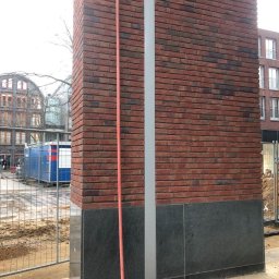 constructionmonter@gmail.com - Fragment elewacji budynku w trakcie budowy z czerwoną cegłą i czarnymi płytami kamiennymi, widoczna rura spustowa i przewody w fundamencie.