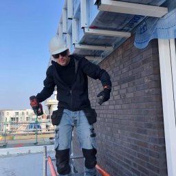 constructionmonter@gmail.com - Pracownik w kasku i okularach słonecznych na rusztowaniu, trzymający wiertarko-wkrętarkę, montuje konstrukcję podsufitki z widoczną izolacją i membraną na tle ceglanej ściany budynku.