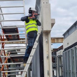 constructionmonter@gmail.com - Pracownik w kasku i kamizelce ochronnej wierci otwór w betonowym słupie na wysokości, stojąc na drabinie opierającej się o rusztowanie.