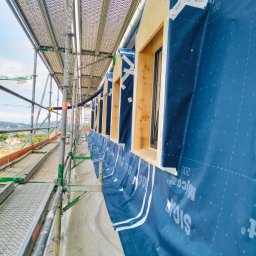 constructionmonter@gmail.com - Budowa domu: widok z rusztowania na przygotowane otwory okienne z drewnianą ramą, zabezpieczone niebieską membraną paroizolacyjną z taśmami uszczelniającymi.