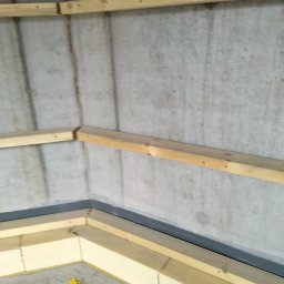 constructionmonter@gmail.com - Konstrukcja szkieletowa z drewnianych belek przy betonowej ścianie, przygotowana do montażu płyty kartonowo-gipsowej, widoczny fragment podłogi z drobnymi odpadami.