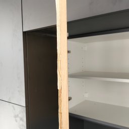 constructionmonter@gmail.com - Przybliżenie drewnianej listwy z widocznymi śladami obróbki, na tle wnętrza szafki kuchennej z półkami.