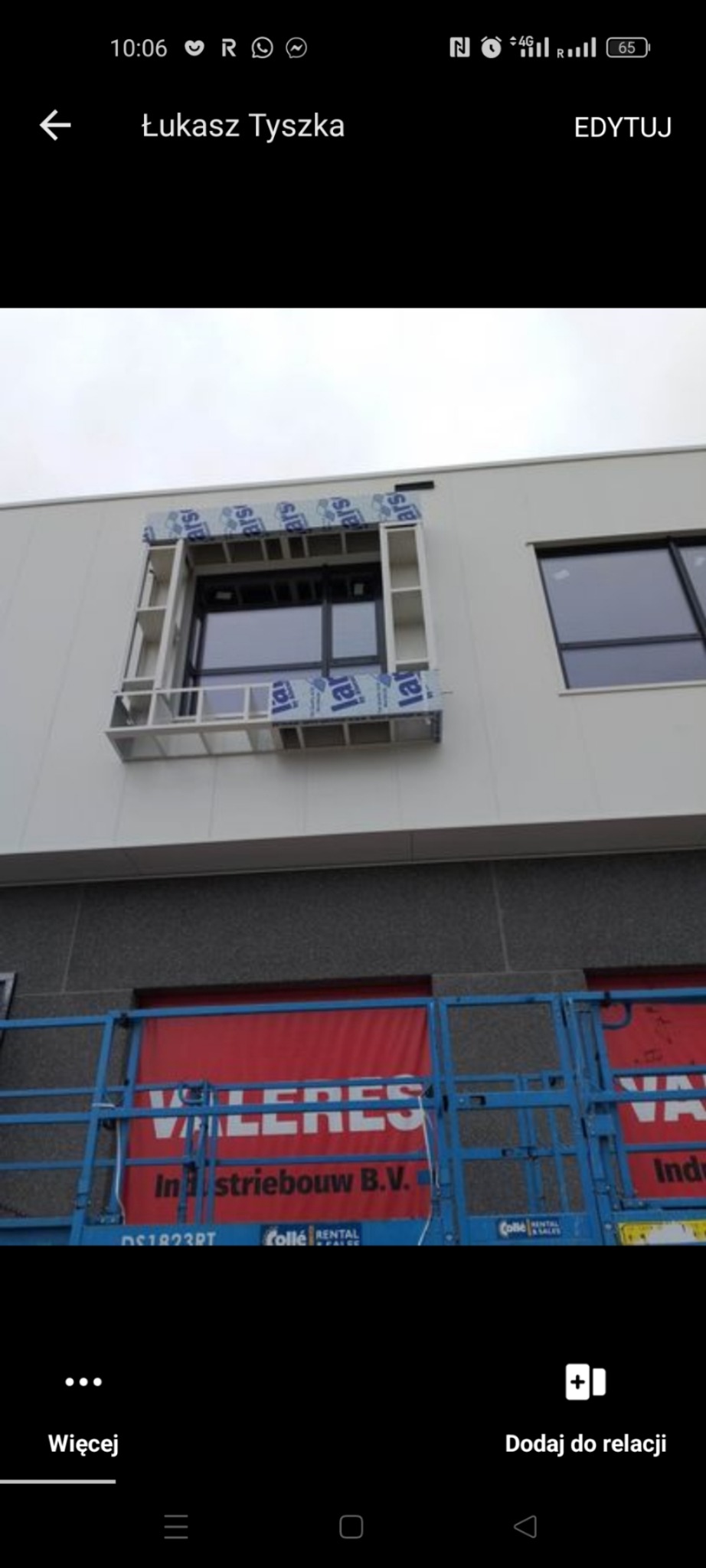 Montaż okna w nowym budynku, widoczna konstrukcja ramy okiennej z zabezpieczeniem folią, na pierwszym planie podnośnik koszowy z banerem reklamowym.