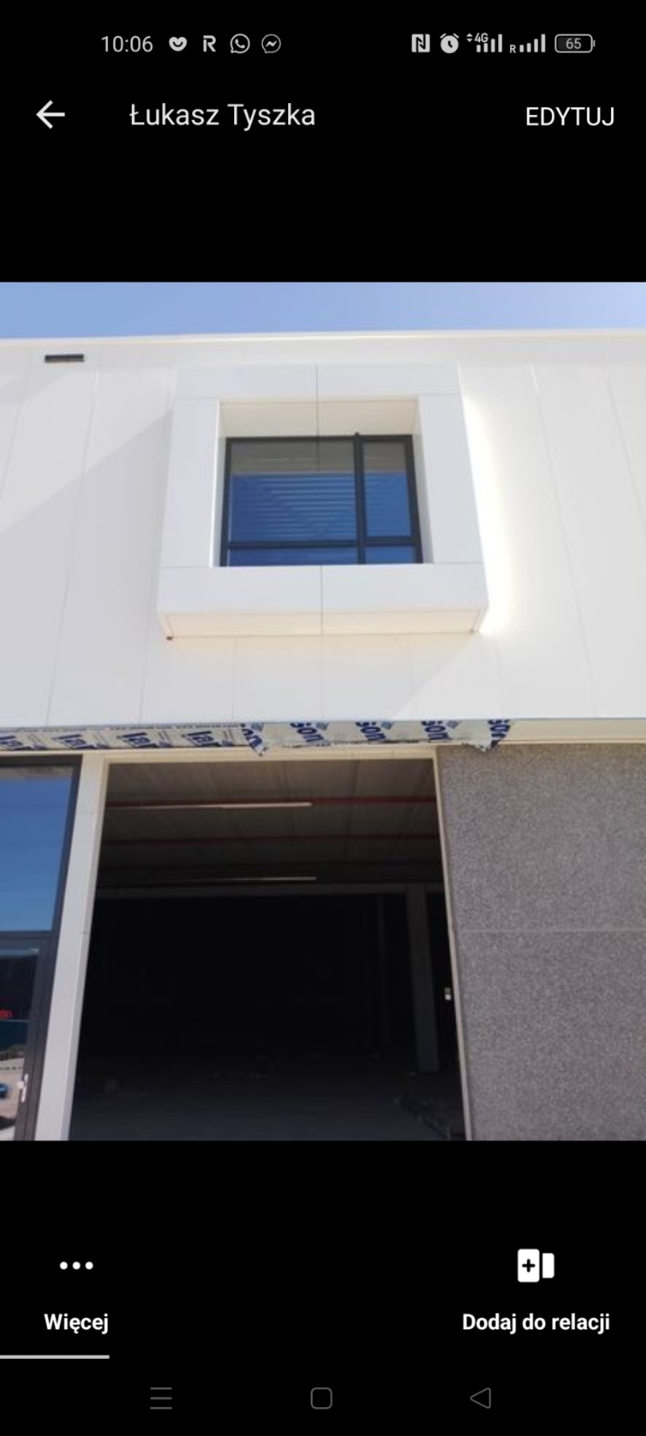 Biała elewacja budynku z oknem w ozdobnej ramie i otwartą bramą wjazdową, zabezpieczoną taśmą malarską na górze.