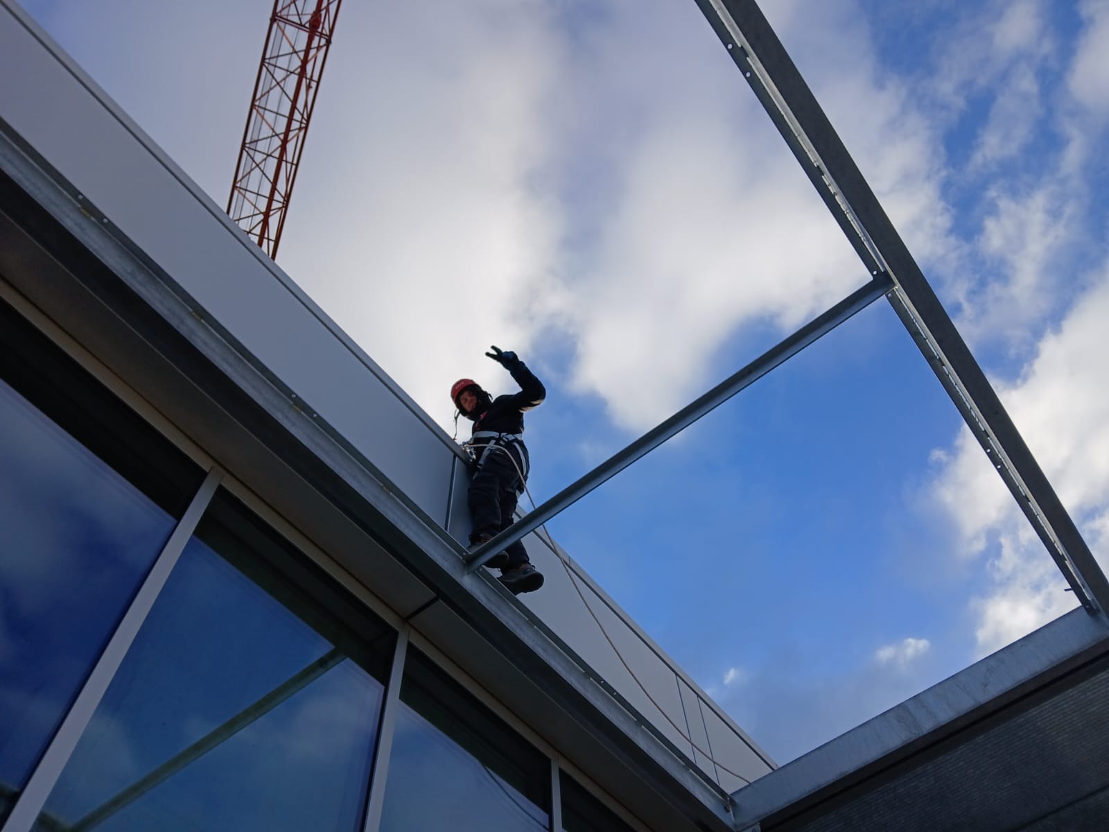 Pracownik w kasku i uprzęży, na wysokości, montuje panele elewacyjne na budynku biurowym, widok z dołu na tle błękitnego nieba i dźwigu.