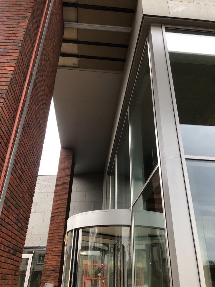 Zewnętrzny widok budynku z ceglaną elewacją i obrotowymi drzwiami, widoczne elementy konstrukcyjne i aluminiowe ramy okienne.