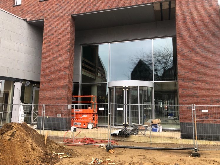 Budynek z cegły z obrotowymi drzwiami wejściowymi w trakcie montażu, widoczny podnośnik koszowy i ogrodzenie tymczasowe.
