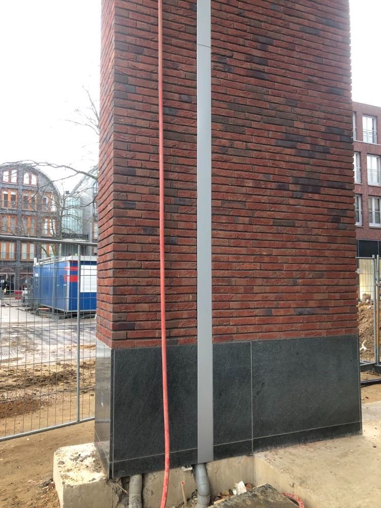 Fragment elewacji budynku w trakcie budowy z czerwoną cegłą i czarnymi płytami kamiennymi, widoczna rura spustowa i przewody w fundamencie.
