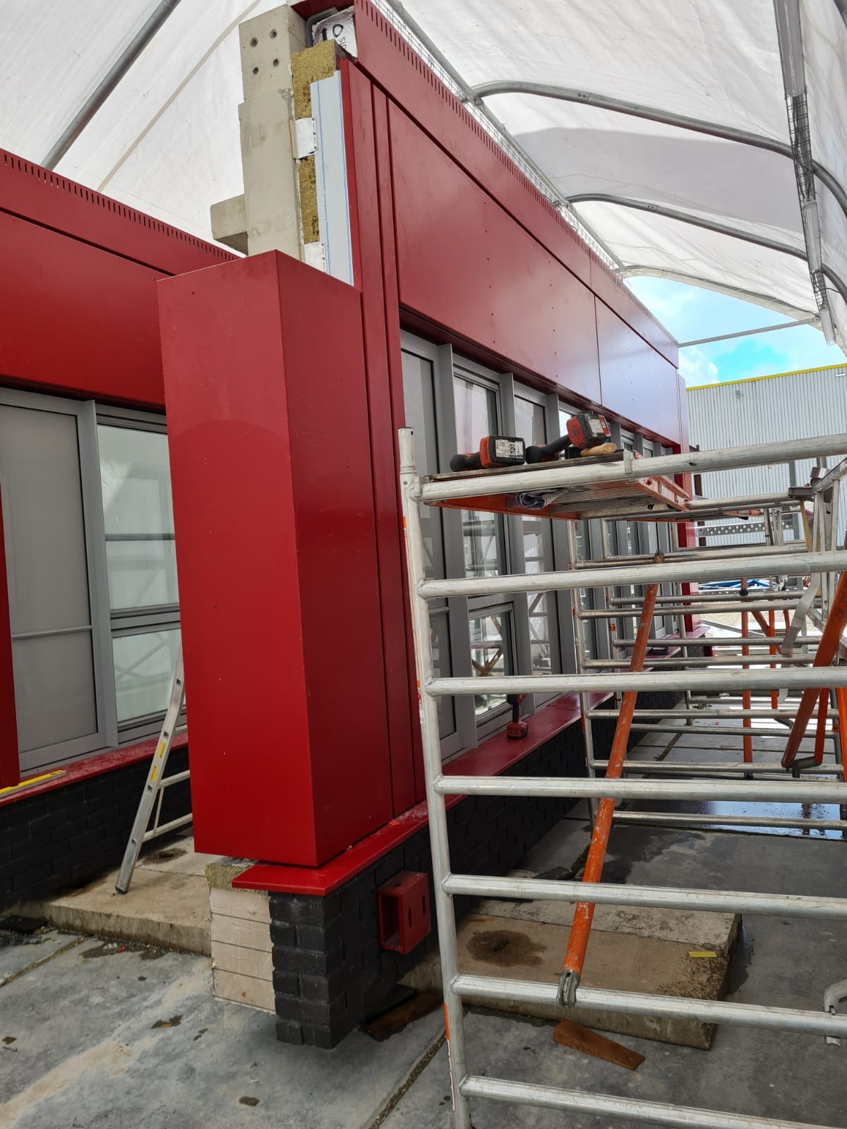 Montaż czerwonej elewacji budynku, widoczne rusztowanie, okna i narzędzia na podeście.