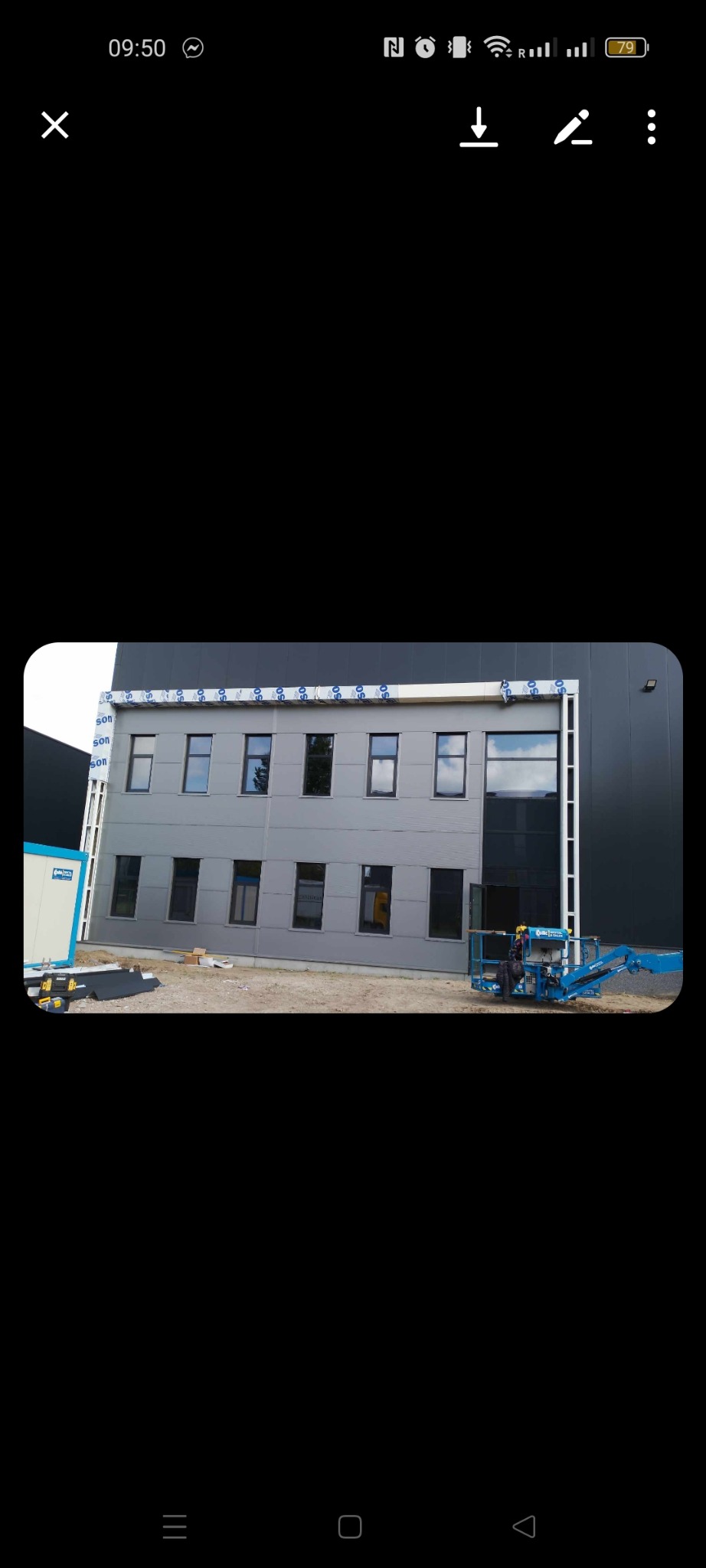 Szary budynek biurowy z czarnymi oknami, widoczna platforma podnośnikowa z pracownikiem w kasku, przygotowania do montażu elewacji.