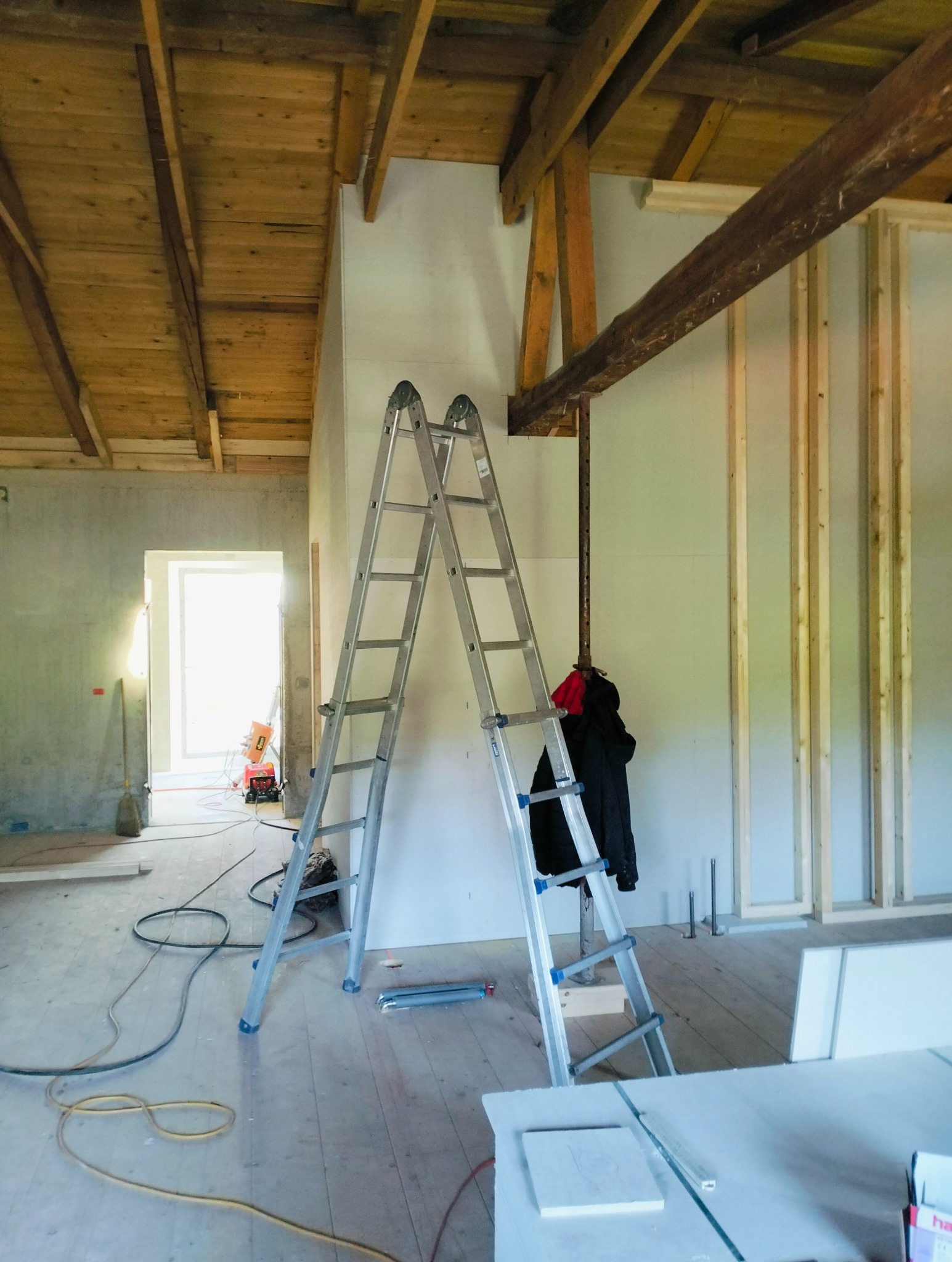 Wnętrze w trakcie remontu: ściany z płyt karton-gips, drewniany strop, metalowa drabina rozstawiona, widoczne kable na podłodze i elementy konstrukcyjne ściany działowej.