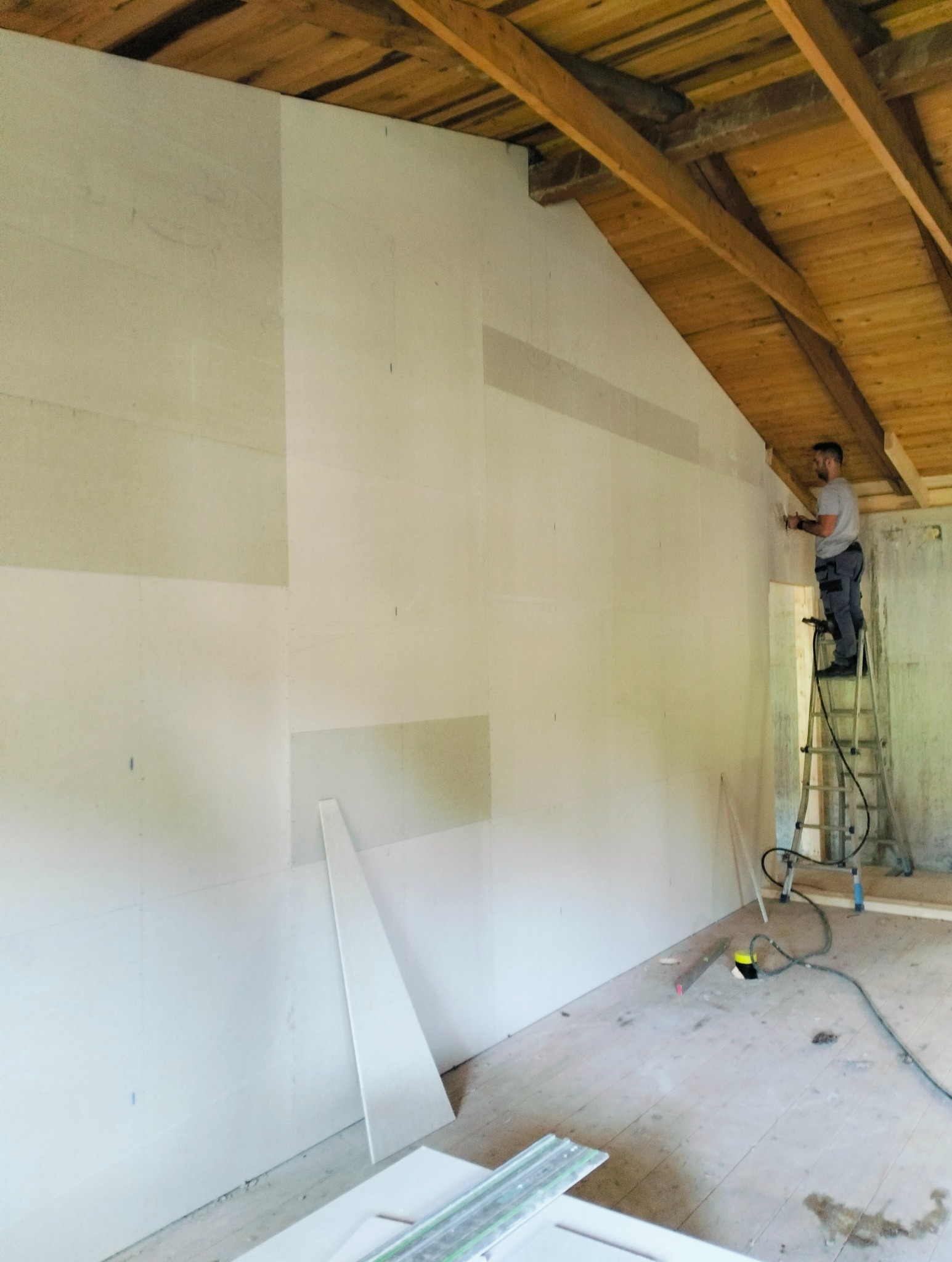 Montaż płyt kartonowo-gipsowych na ścianie poddasza, widoczny pracownik na drabinie, częściowo zamontowane płyty i drewniana konstrukcja dachu.