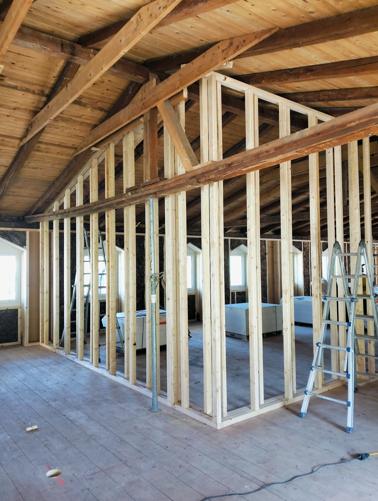 Konstrukcja ścian działowych z drewnianych belek na poddaszu z drewnianą boazerią, widoczne rusztowania i materiały budowlane w tle.