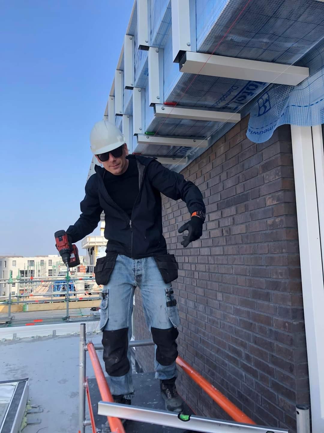 Pracownik w kasku i okularach słonecznych na rusztowaniu, trzymający wiertarko-wkrętarkę, montuje konstrukcję podsufitki z widoczną izolacją i membraną na tle ceglanej ściany budynku.