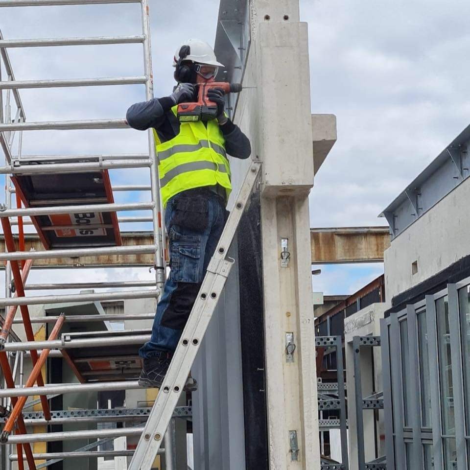 Pracownik w kasku i kamizelce ochronnej wierci otwór w betonowym słupie na wysokości, stojąc na drabinie opierającej się o rusztowanie.