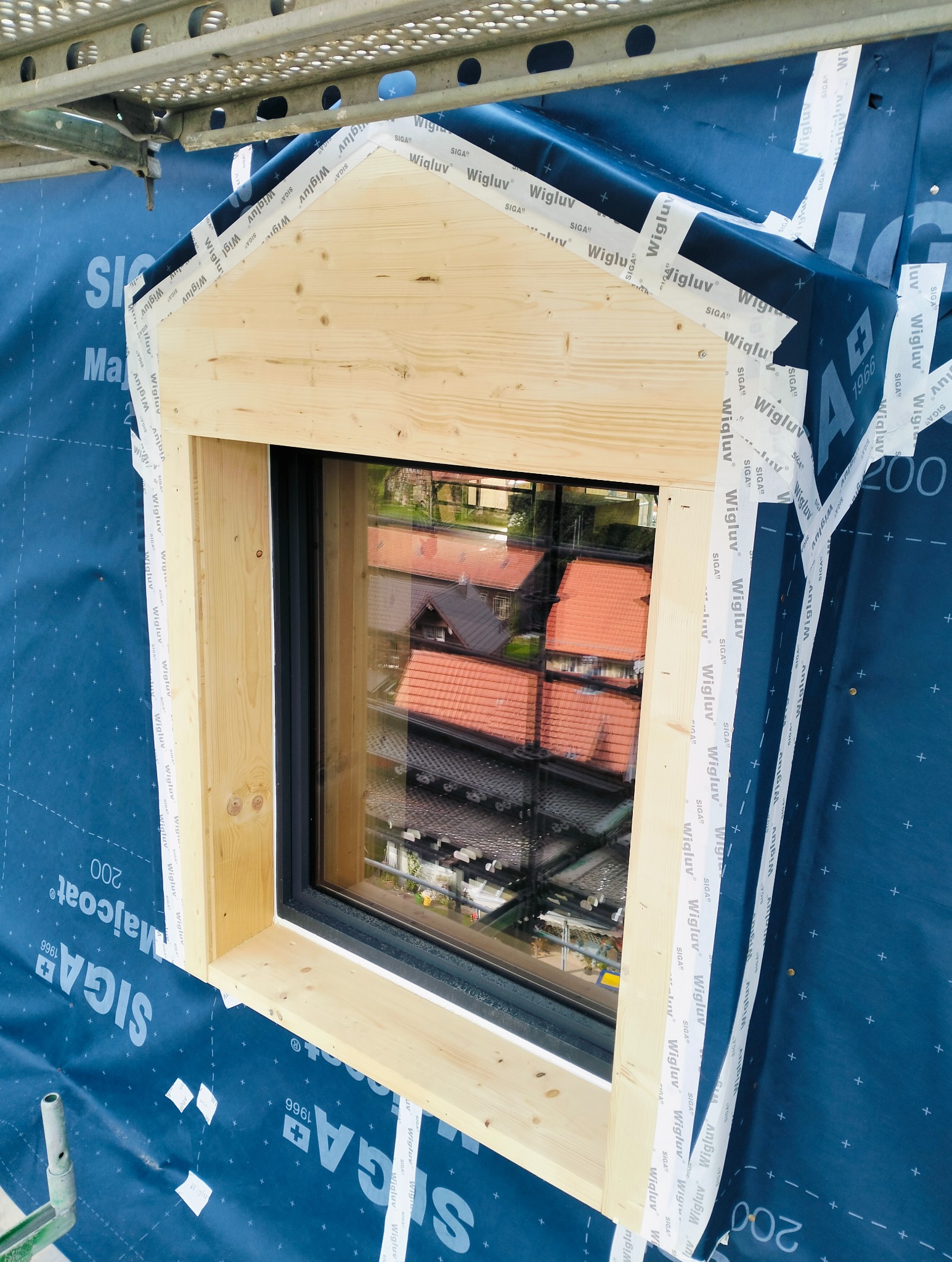 Surowe drewno ramy okiennej, widok z zewnątrz, zabezpieczone folią i taśmą uszczelniającą na tle niebieskiej membrany dachowej, widok na dachy sąsiednich budynków odbijający się w szybie.