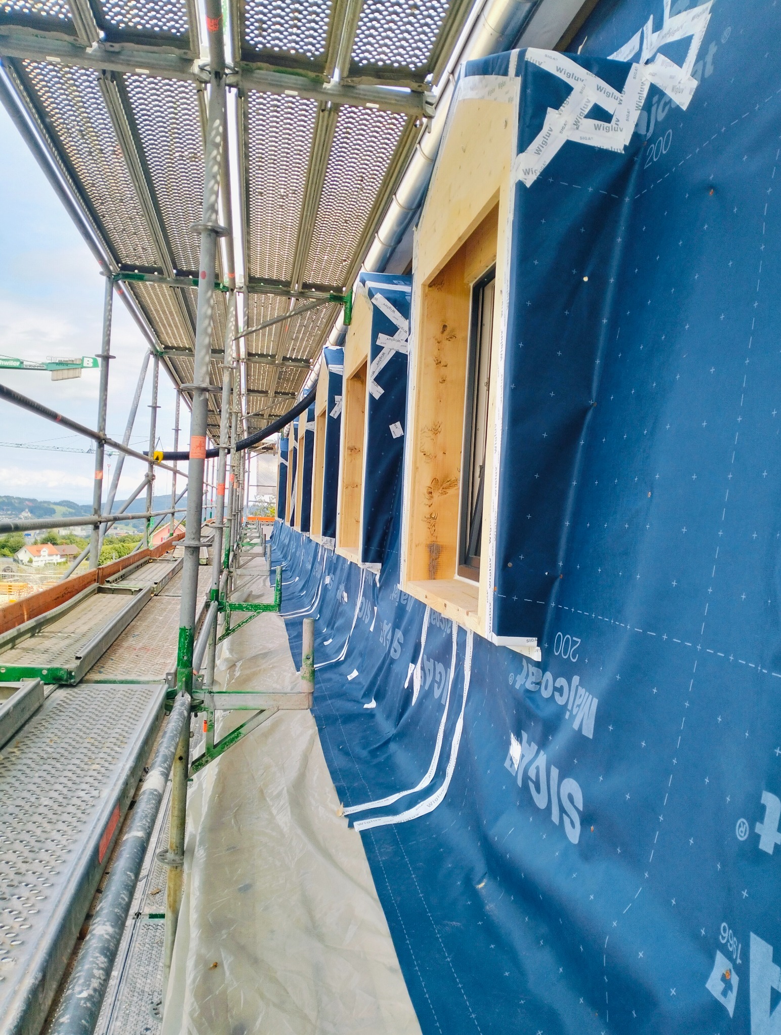 Budowa domu: widok z rusztowania na przygotowane otwory okienne z drewnianą ramą, zabezpieczone niebieską membraną paroizolacyjną z taśmami uszczelniającymi.