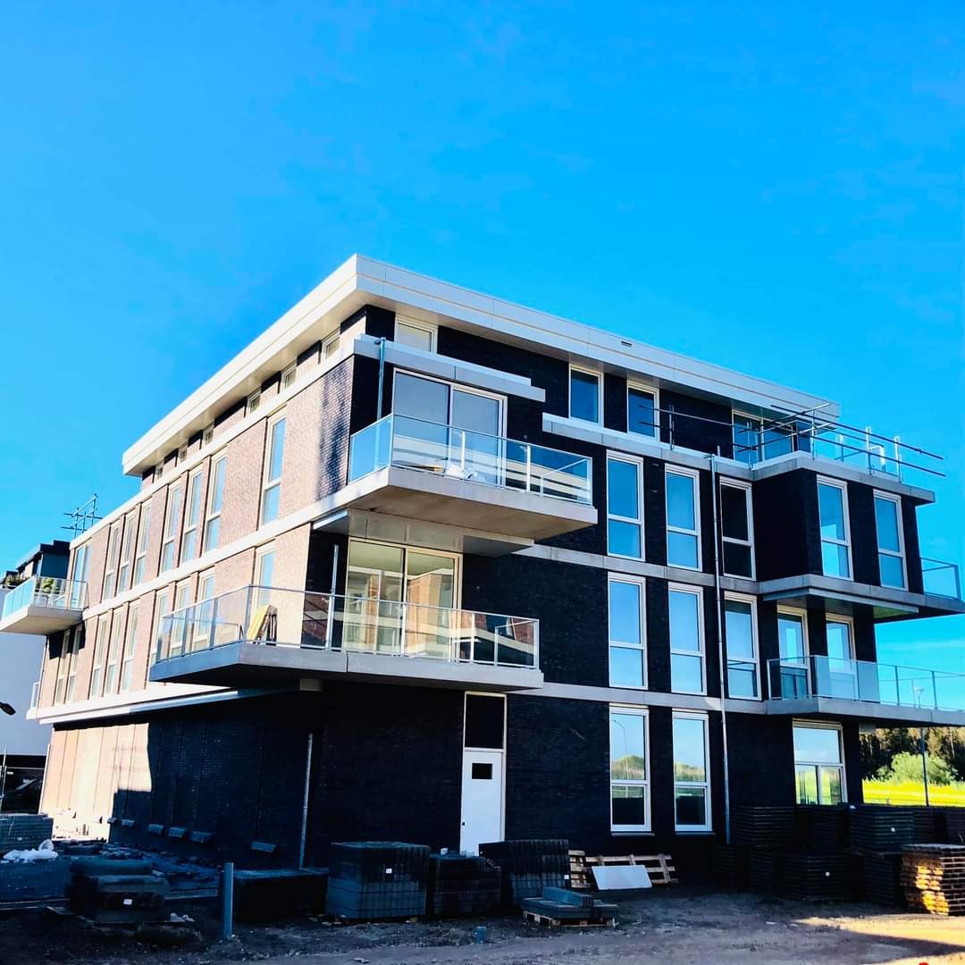 Nowoczesny, trzypiętrowy budynek z ciemnej cegły z balkonami z przezroczystymi balustradami, widoczny w trakcie budowy z paletami materiałów budowlanych na pierwszym planie.