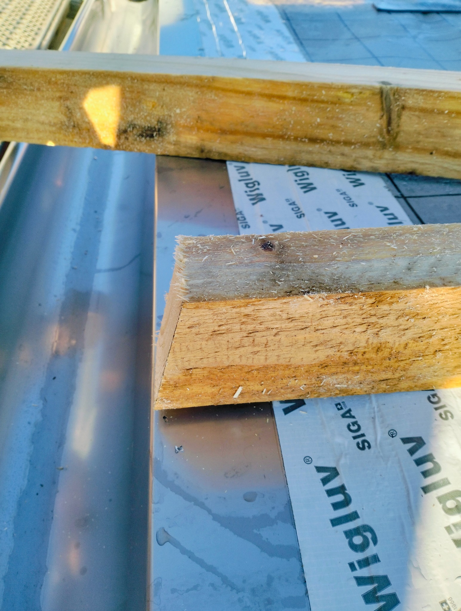 Fragment konstrukcji dachu z widocznymi elementami drewnianymi, srebrną blachą i membraną dachową z nadrukiem Wigluv, uchwycony z bliskiej perspektywy.