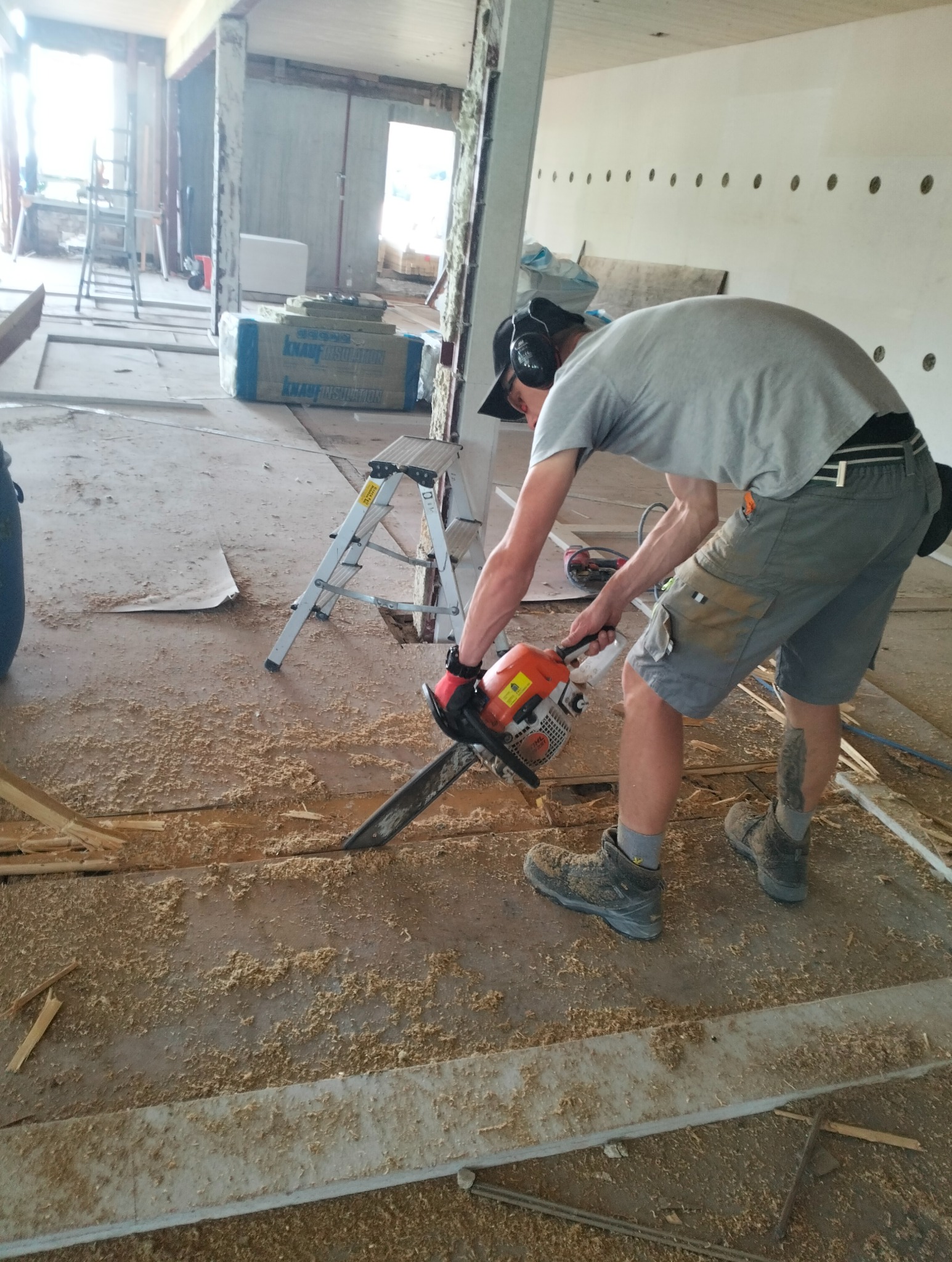 Mężczyzna w ochronnikach słuchu tnie piłą spalinową podłogę w trakcie remontu, w tle widoczne materiały budowlane i drabina.
