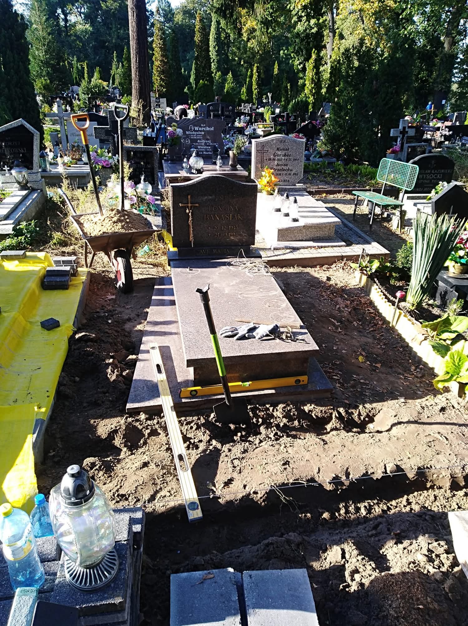 Montaż nagrobka na cmentarzu: poziomica, łopata, taczka z ziemią. Przygotowanie terenu pod pomnik, narzędzia pracy. Prace kamieniarskie w toku, widok z góry.