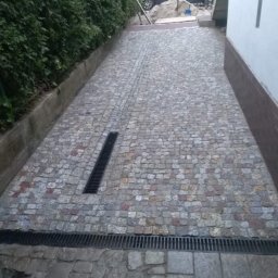 Wolfheart prace brukarskie - Ułożona kostka granitowa tworząca ścieżkę z odpływami liniowymi. W tle zieleń i częściowo widoczny samochód. Kąt ujęcia z dołu, podkreślający perspektywę.