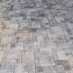 Wolfheart prace brukarskie - Ułożona szara kostka brukowa w nieregularny wzór, widok z góry. Ciemne fugi podkreślają kształt kostek. W tle brama garażowa i częściowo widoczna sylwetka osoby.