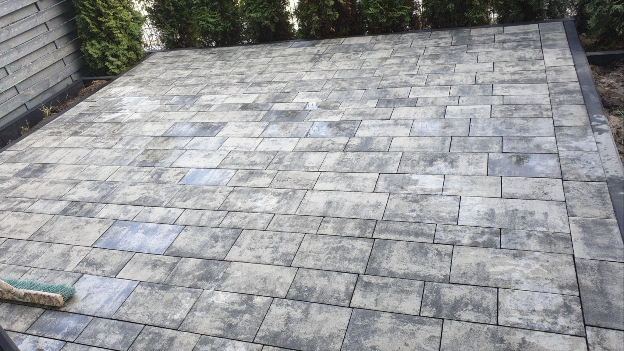 Ułożona kostka brukowa w odcieniach szarości, tworząca równą powierzchnię tarasu lub podjazdu, z widoczną szczotką do zamiatania i zielenią w tle.