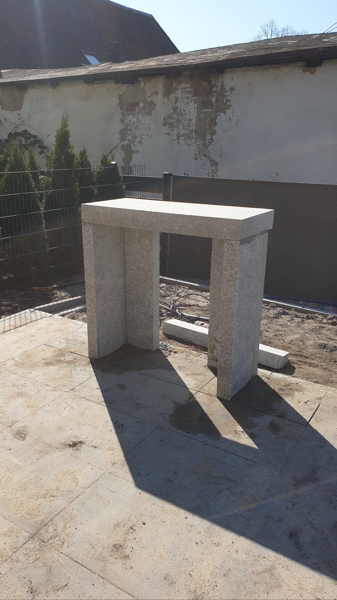 Granitowa konstrukcja w kształcie portalu na świeżo ułożonym bruku, w tle ogrodzenie i budynek z jasną elewacją. Słoneczny dzień.